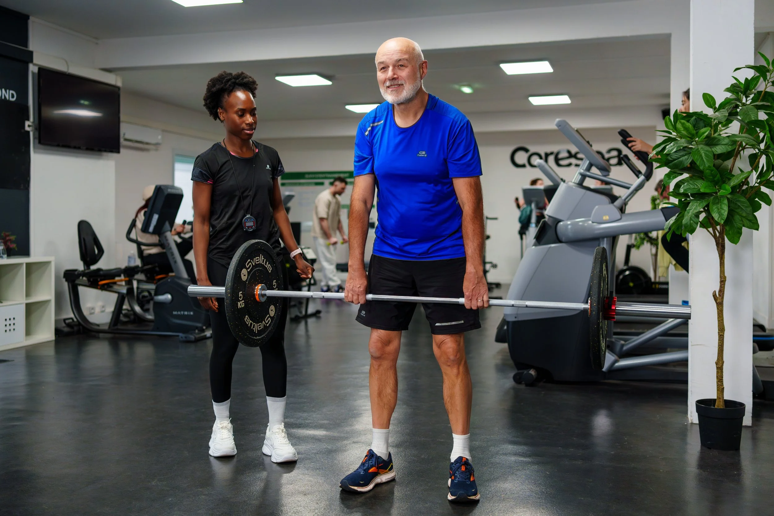 Un homme âgé en train de soulever une barre avec un poids dans une salle de sport, assisté par une entraîneuse ou une accompagnatrice.
