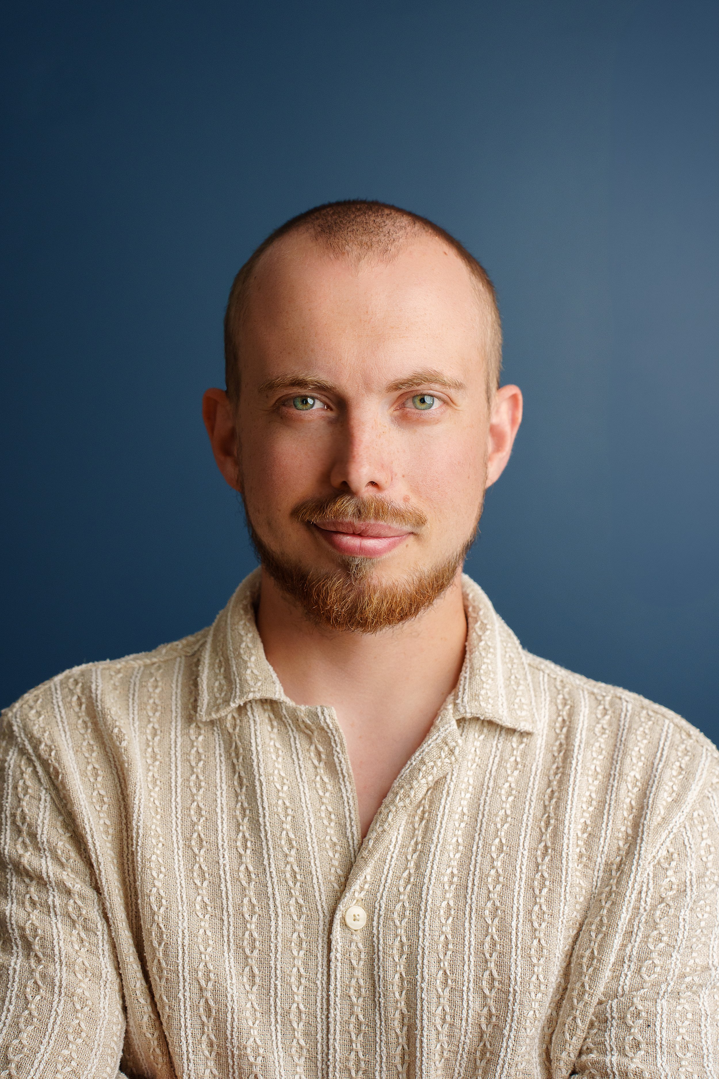 Portrait d'un homme aux cheveux ras, barbe légère, portant une chemise beige à rayures, contre un fond bleu