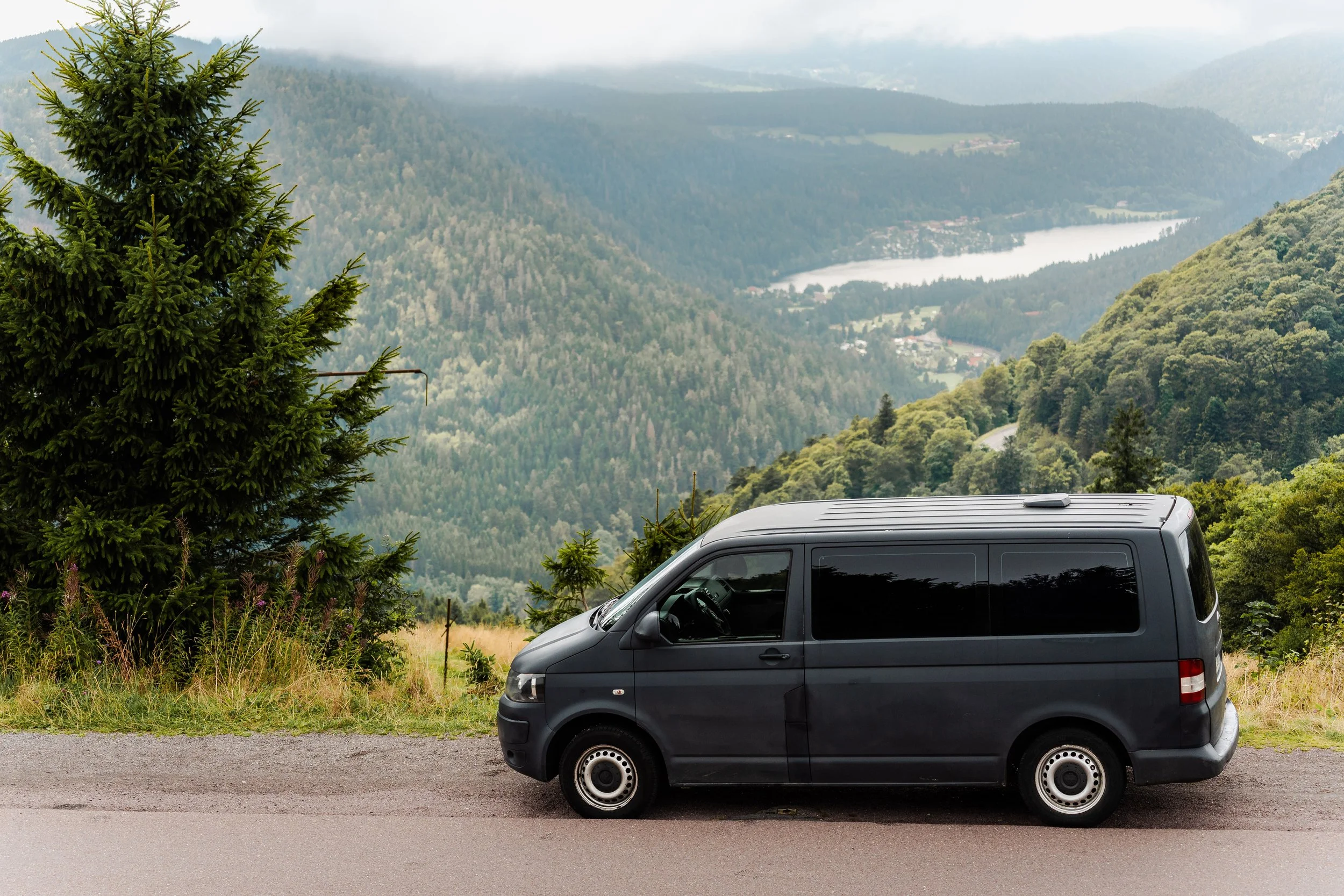 Véhicule utilitaire noir garé sur le bord d'une route de montagne avec un paysage de forêt, de vallée, et de lac en arrière-plan.