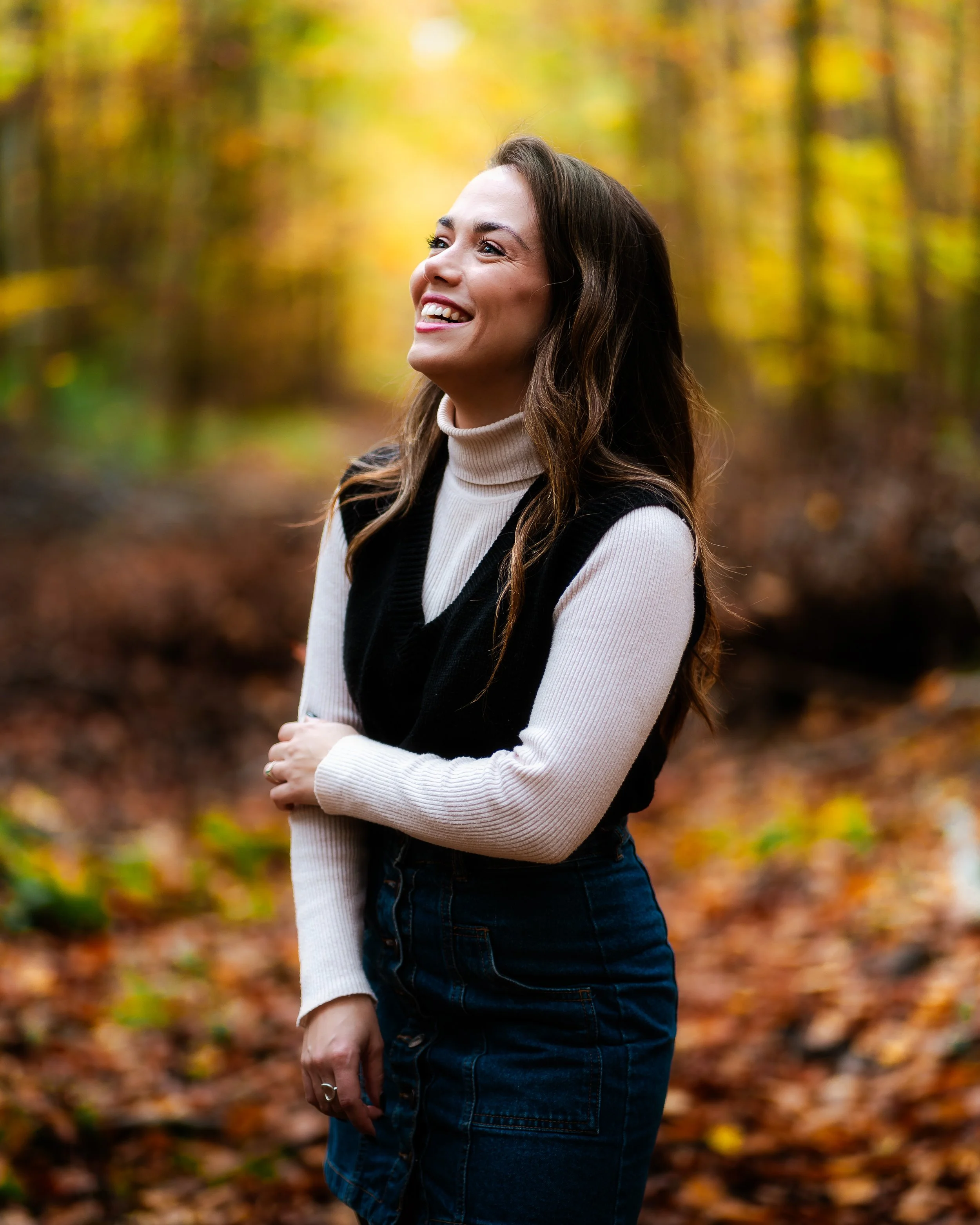 Femme souriante dans un bois d'automne, portant un pull beige, un gilet noir et une jupe en jean.
