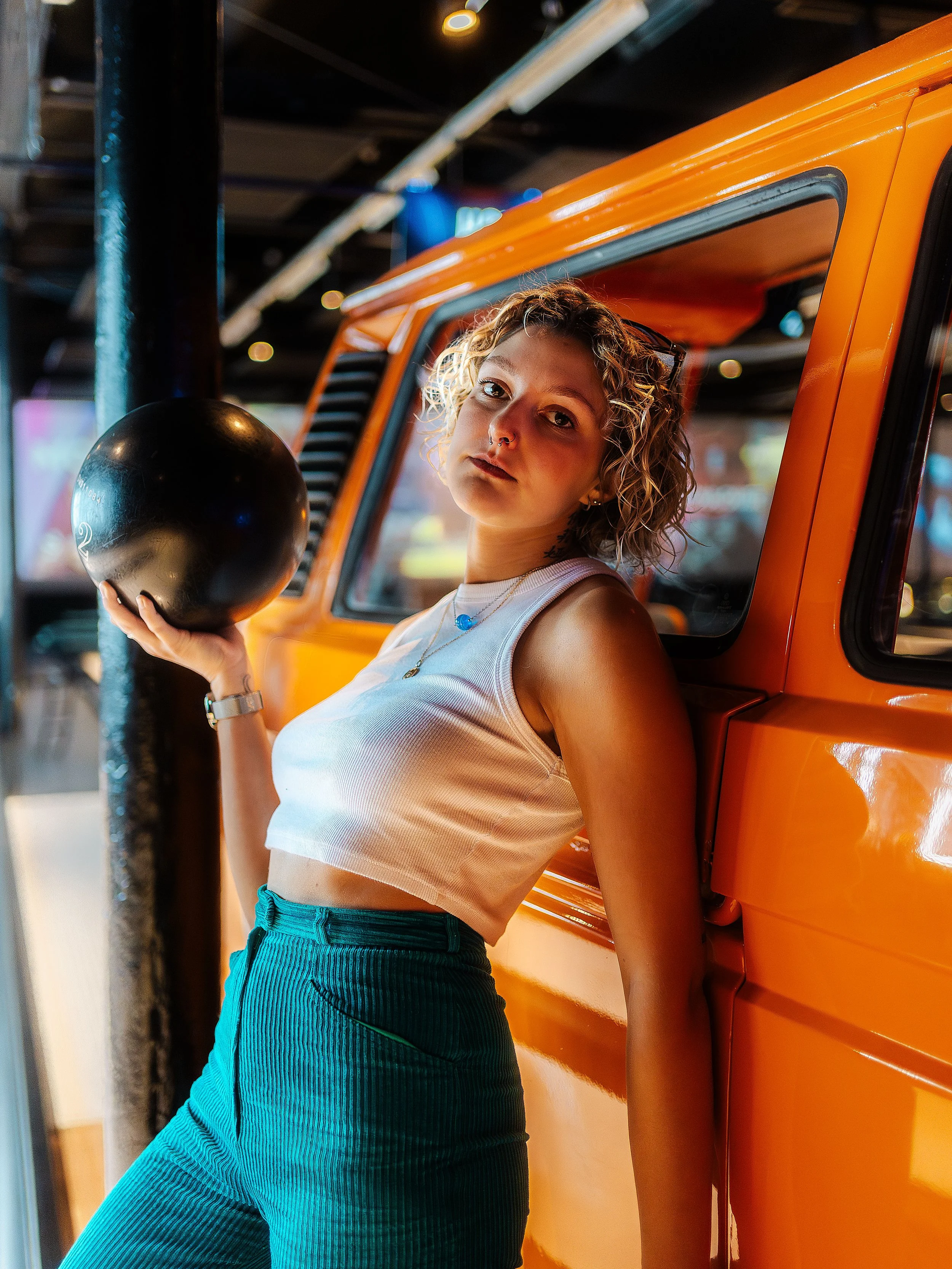 Une jeune femme aux cheveux bouclés, portant un débardeur blanc et un pantalon rayé bleu-vert, tient une boule noire dans une main, appuyée contre un véhicule orange dans un environnement intérieur.