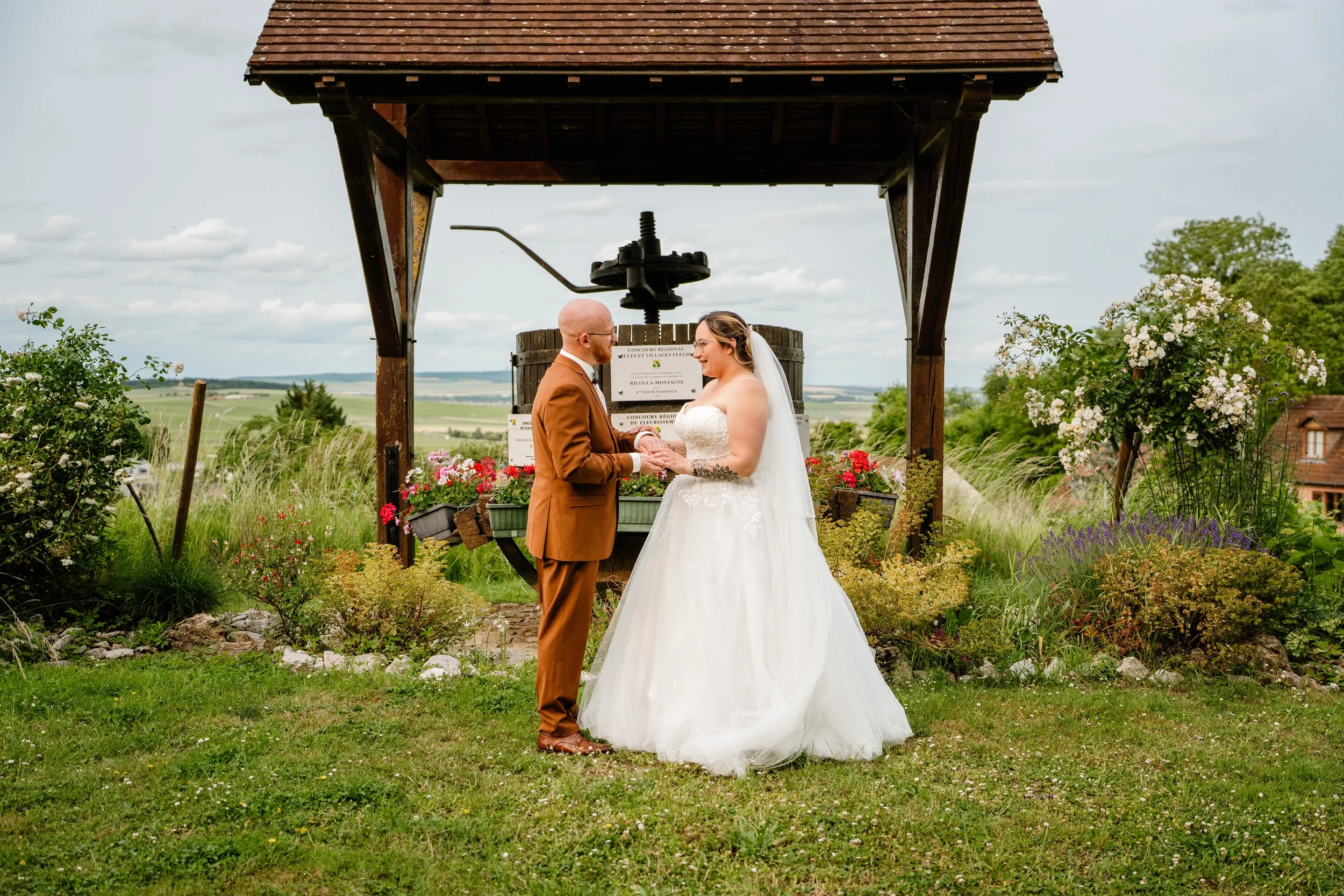 Un couple de mariés échangeant leurs vœux lors de leur mariage en plein air, devant un moulin à eau en bois, entouré de fleurs et de verdure, avec un paysage rural en arrière-plan.