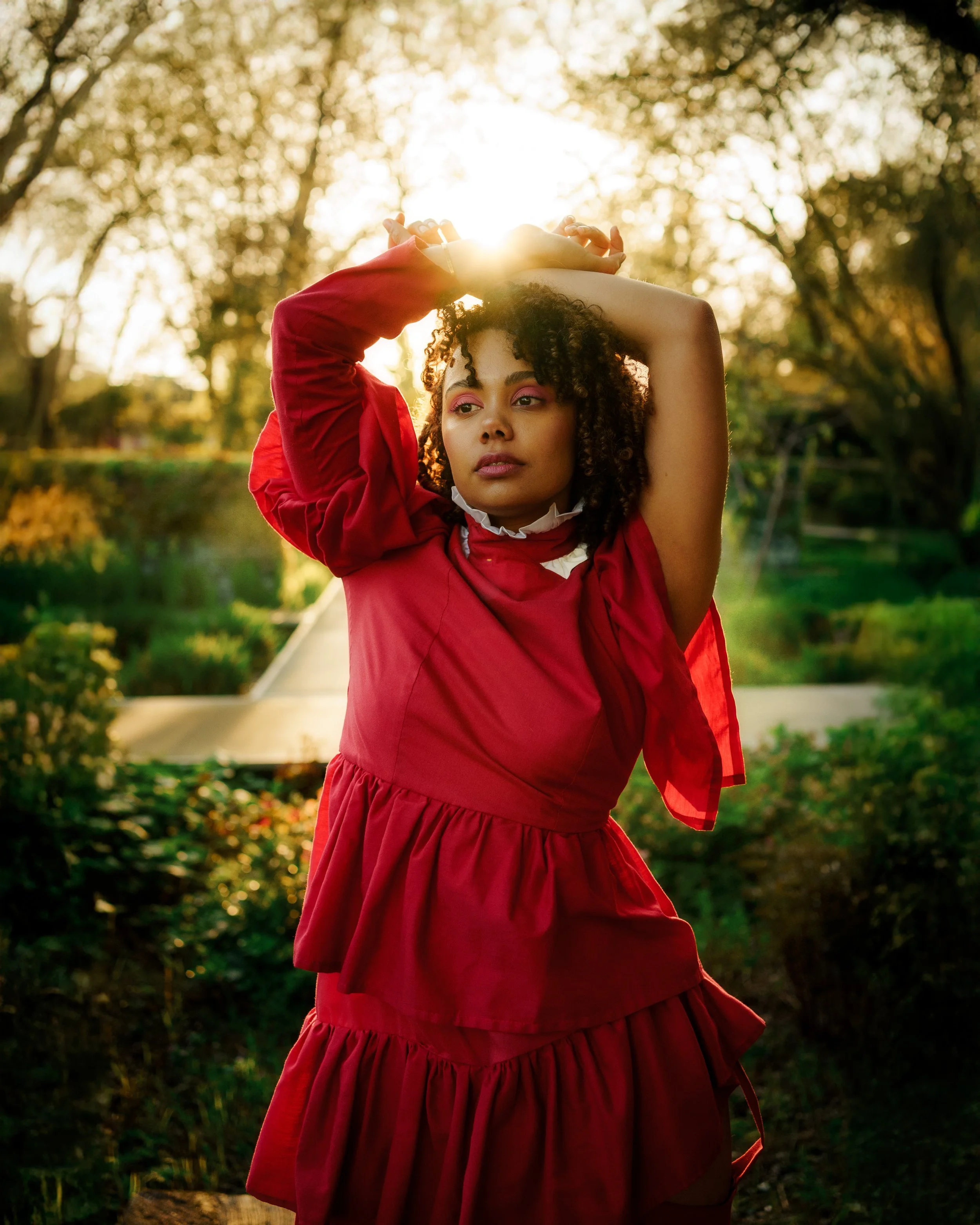 Une femme en robe rouge pose avec les bras levés, tenant sa tête, dans un parc en été avec le soleil couchant en arrière-plan.