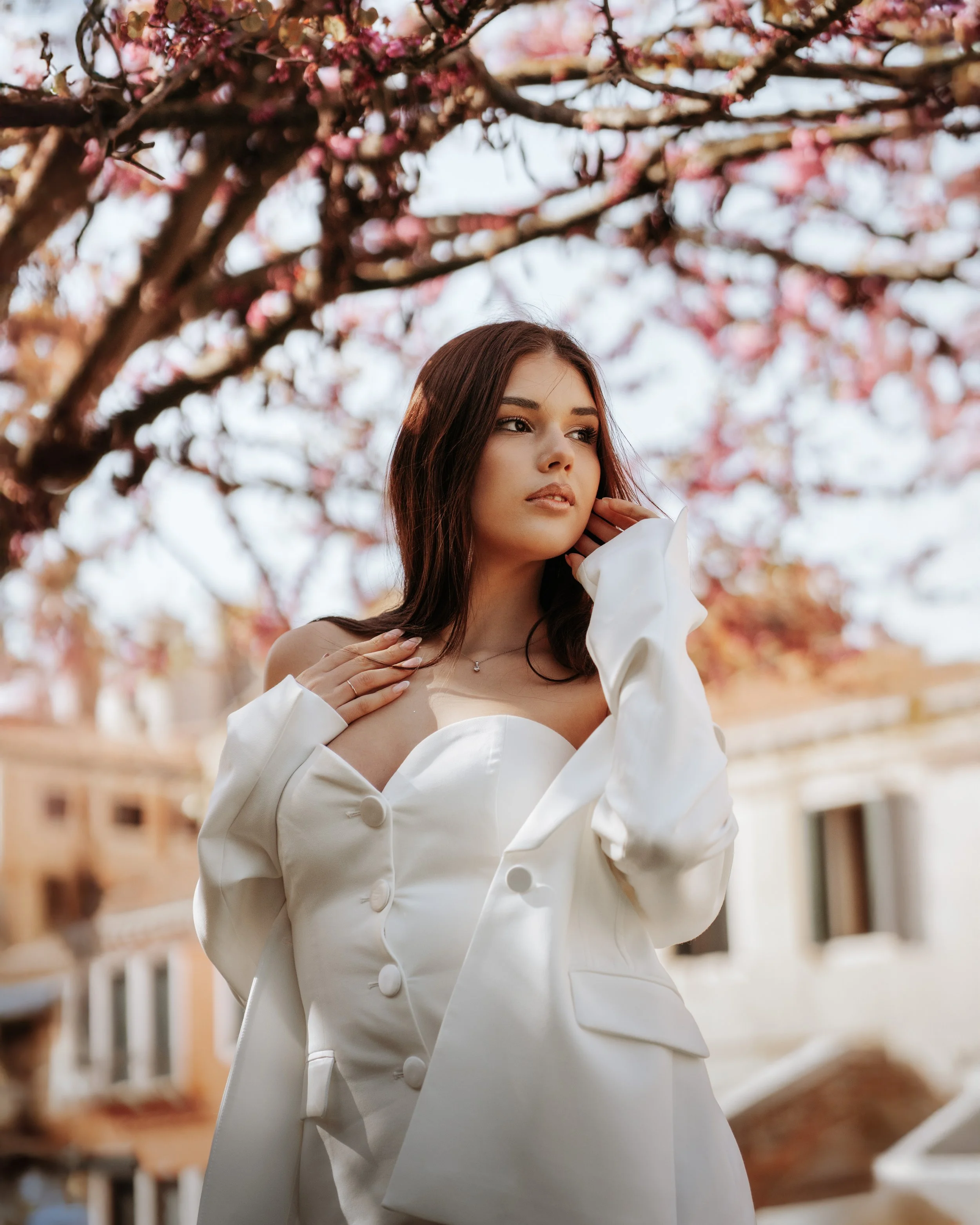 Une jeune femme avec des cheveux bruns, portant une robe blanche et une veste blanche, se tient sous un arbre en fleurs roses, devant un bâtiment résidentiel.