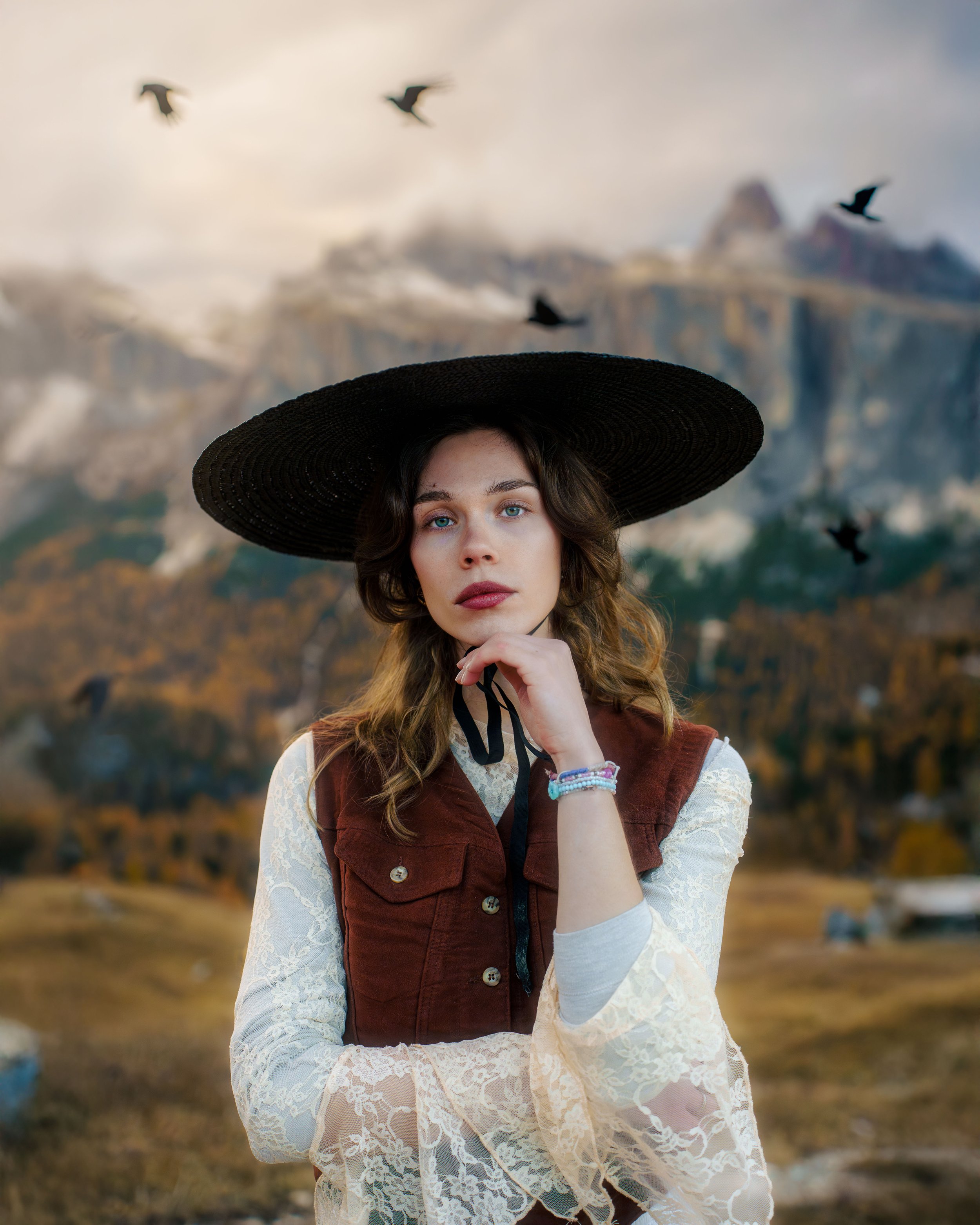 Une jeune femme porte un grand chapeau noir, un gilet marron et un haut en dentelle, se tenant dans un paysage de montagne avec des oiseaux en vol en arrière-plan.