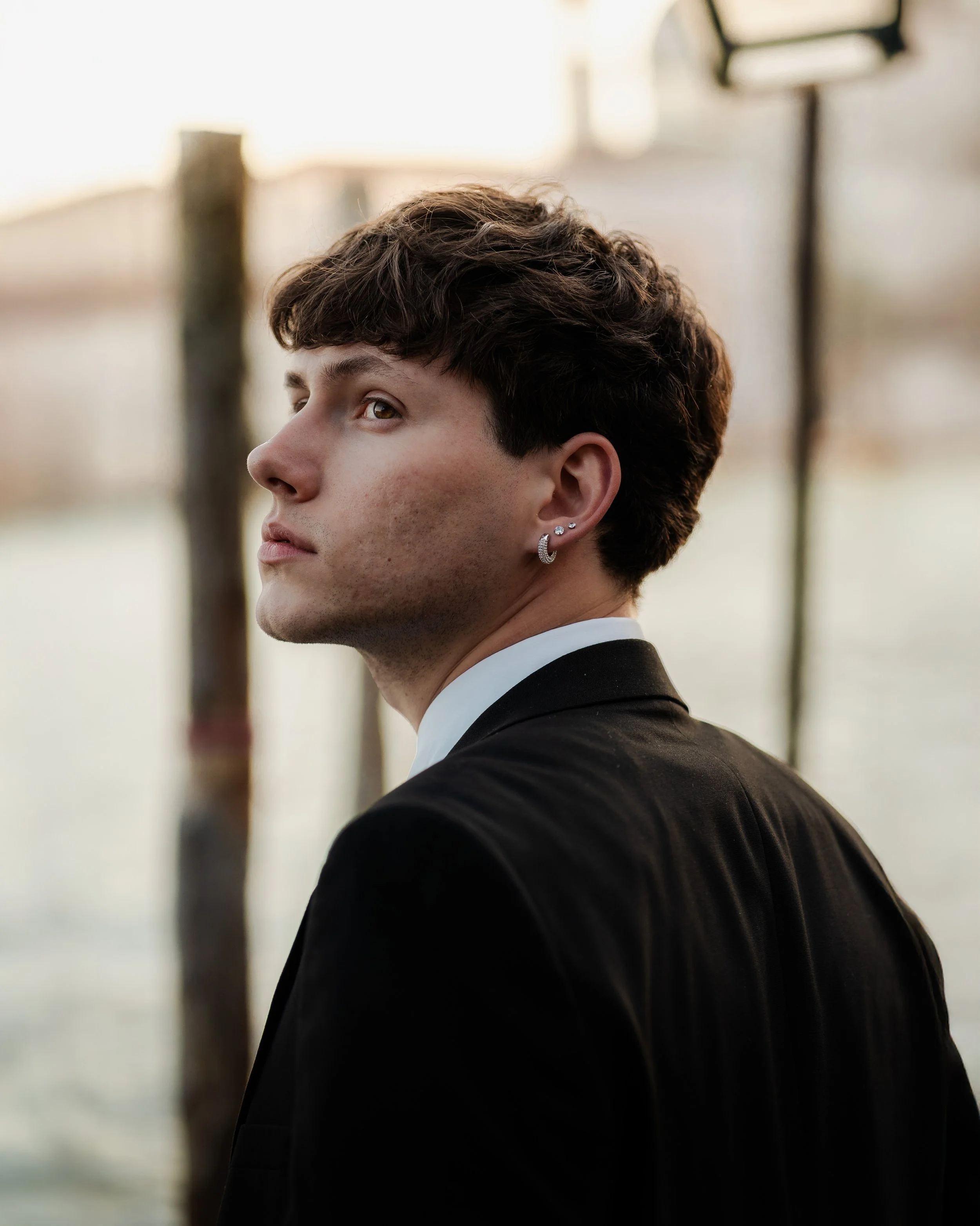 Jeune homme avec boucles d'oreilles portant un costume noire, regardant vers la gauche, en extérieur près de l'eau au coucher du soleil.