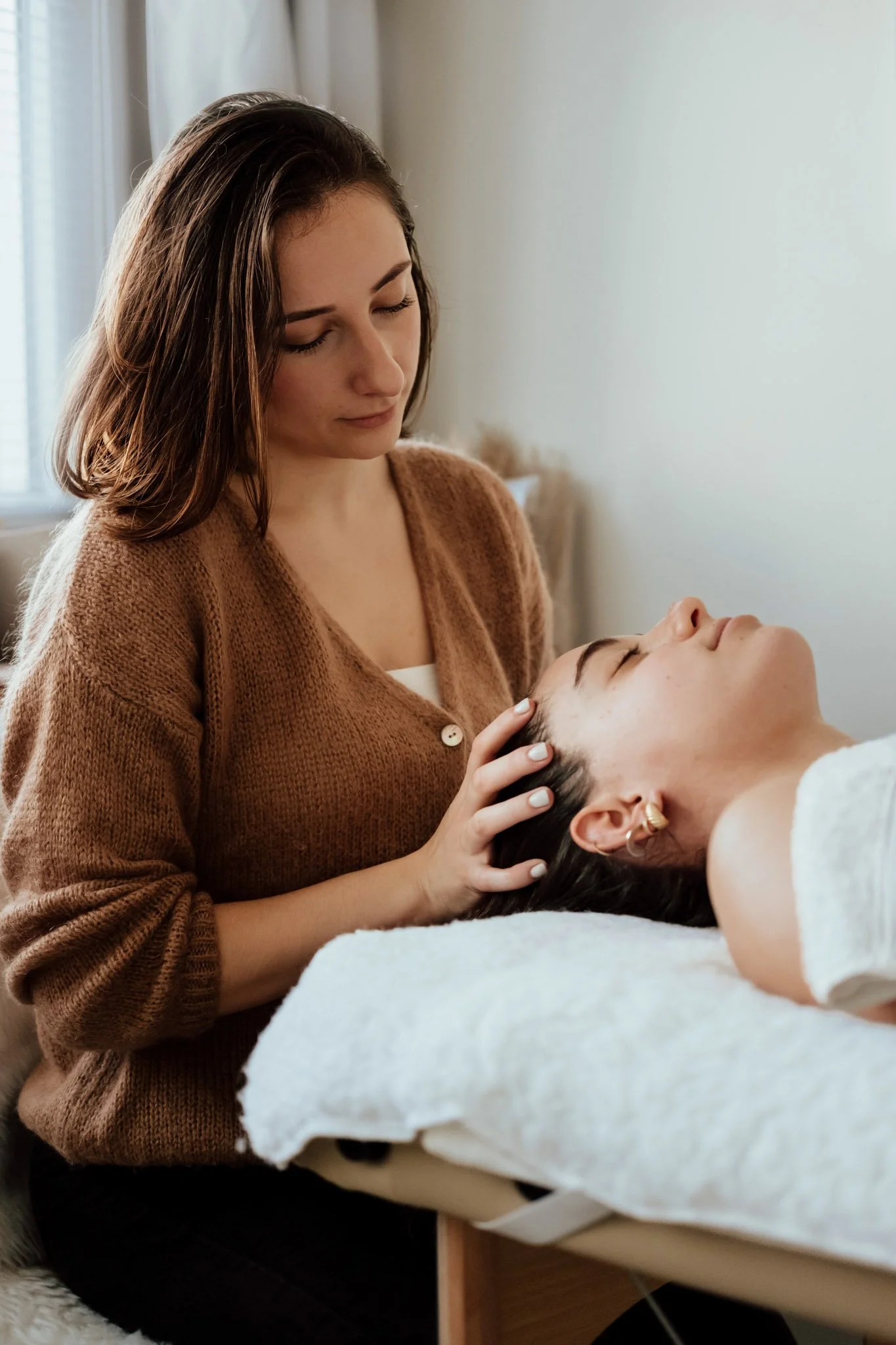 Une femme donne un massage crânien à une autre femme allongée sur une table de massage dans une pièce lumineuse.