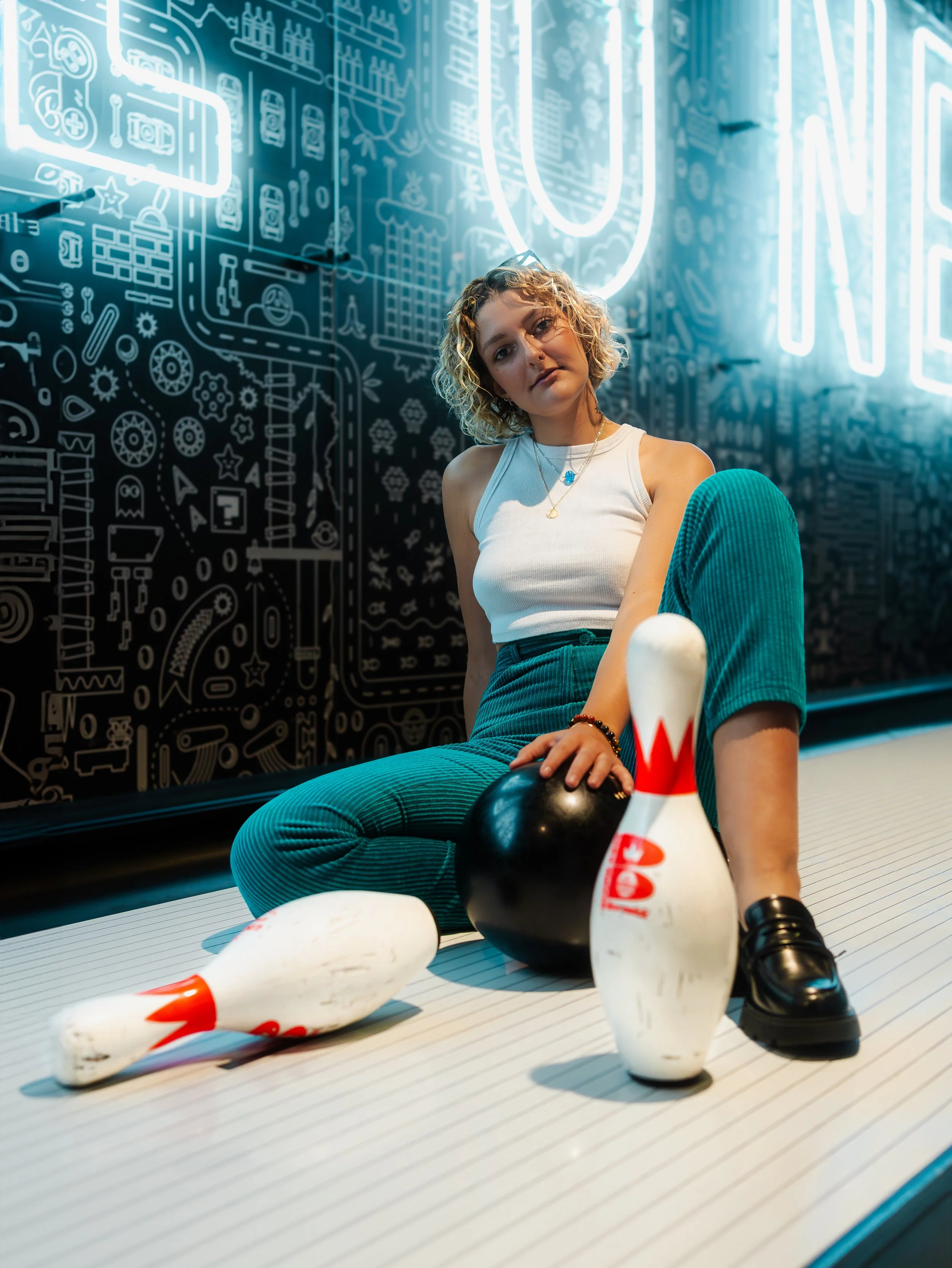 Jeune femme avec cheveux bouclés, portant un débardeur blanc, assise sur le sol à côté de quilles de bowling, dans une salle de bowling avec un décor noir et des lumières néon.