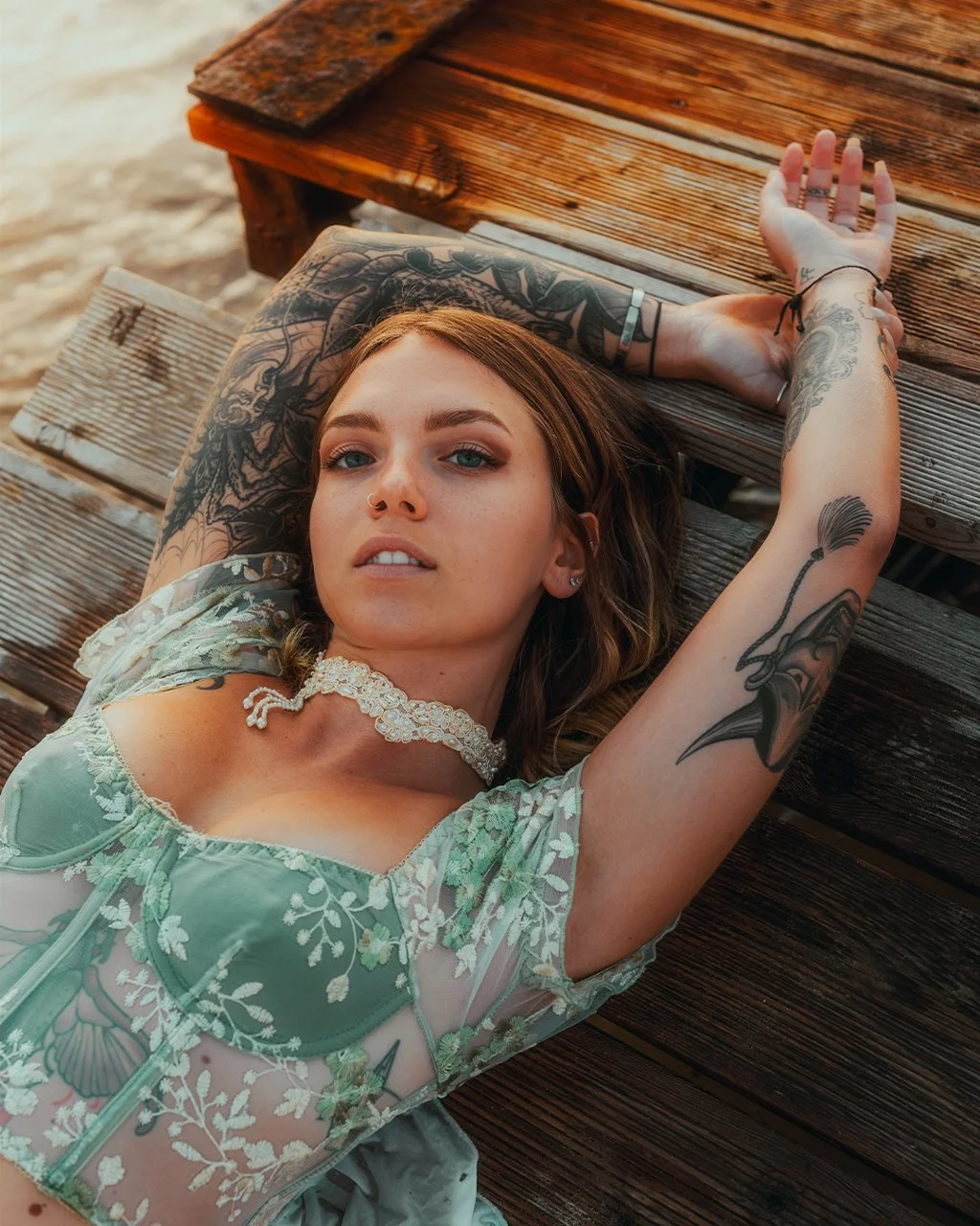 Jeune femme aux tatouages allongée sur un banc en bois, portant une robe en dentelle verte et un collier, regardant vers l'objectif.