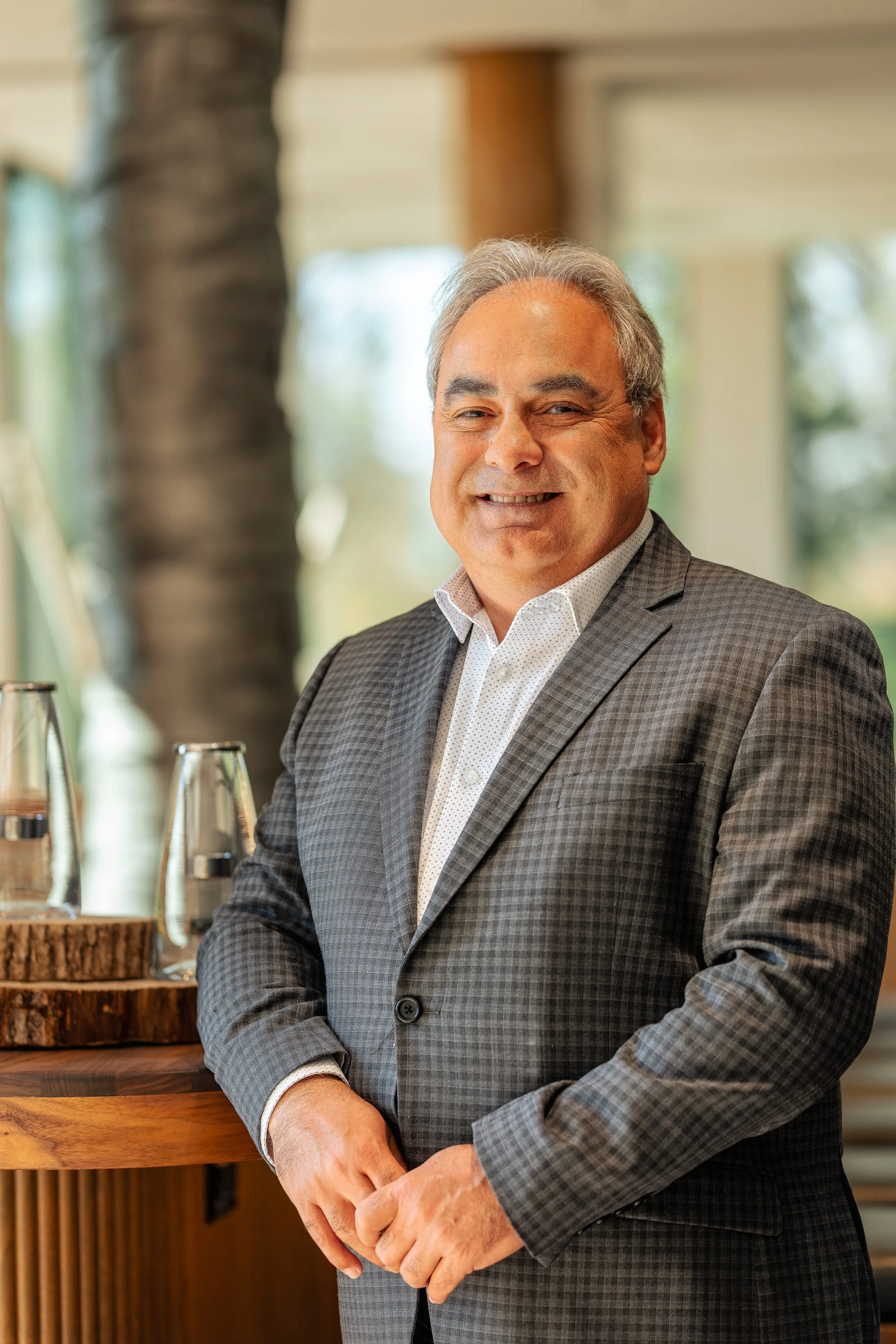 Homme d'âge moyen en costume souriant, debout à côté d'une table en bois avec des verres à eau et des décorations en bois.