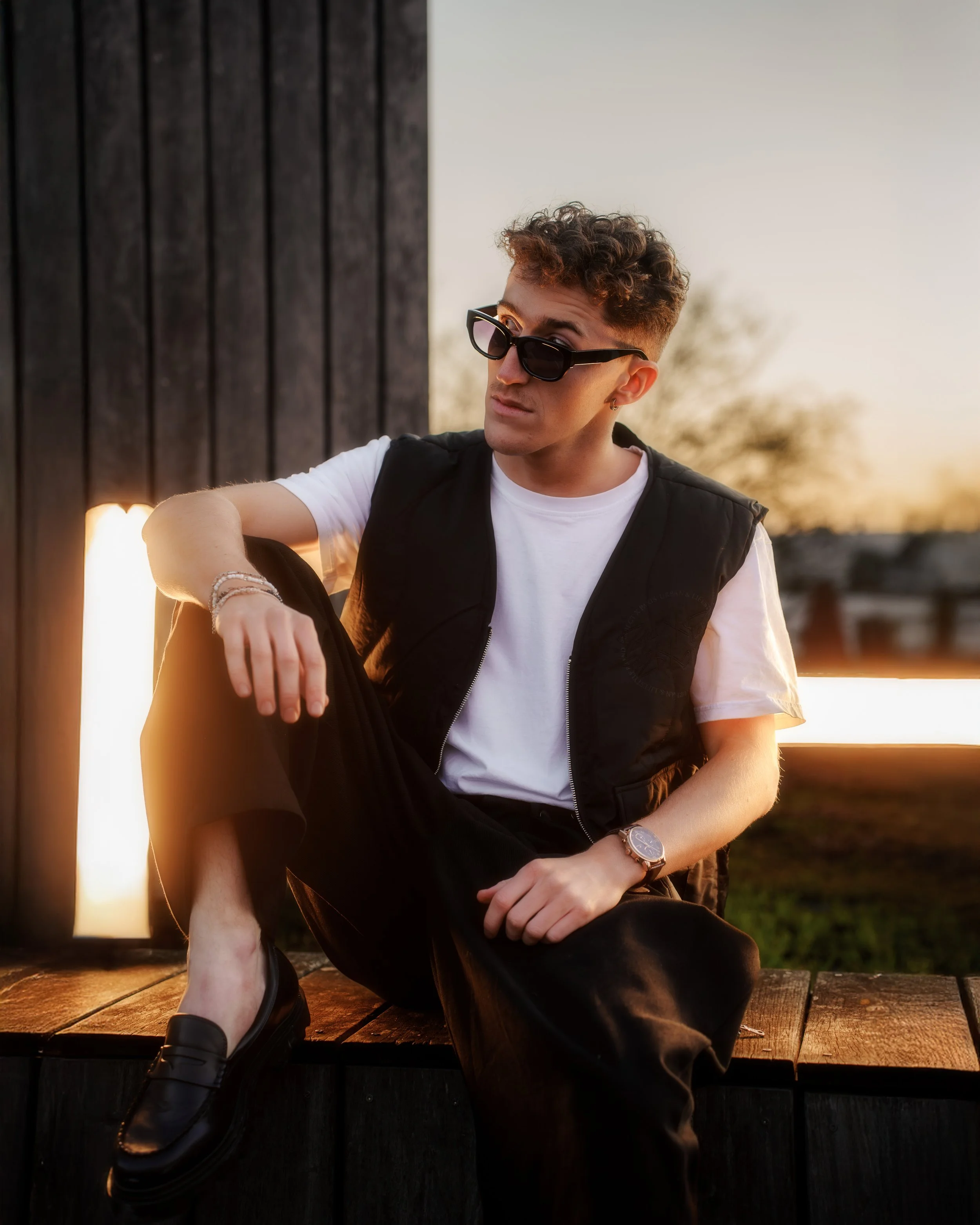 Jeune homme avec des lunettes de soleil, portant un t-shirt blanc et un gilet noir, assis sur une plateforme en bois lors d'un coucher de soleil, avec un fond de ciel clair et arbres squelettiques.