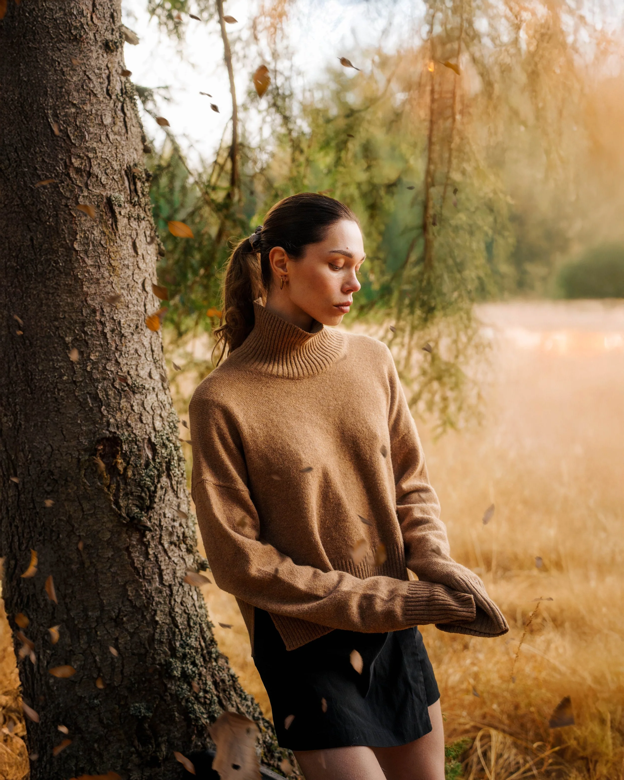 Jeune femme en pull marron, les yeux fermés, debout derrière un arbre en automne avec des feuilles tombées.