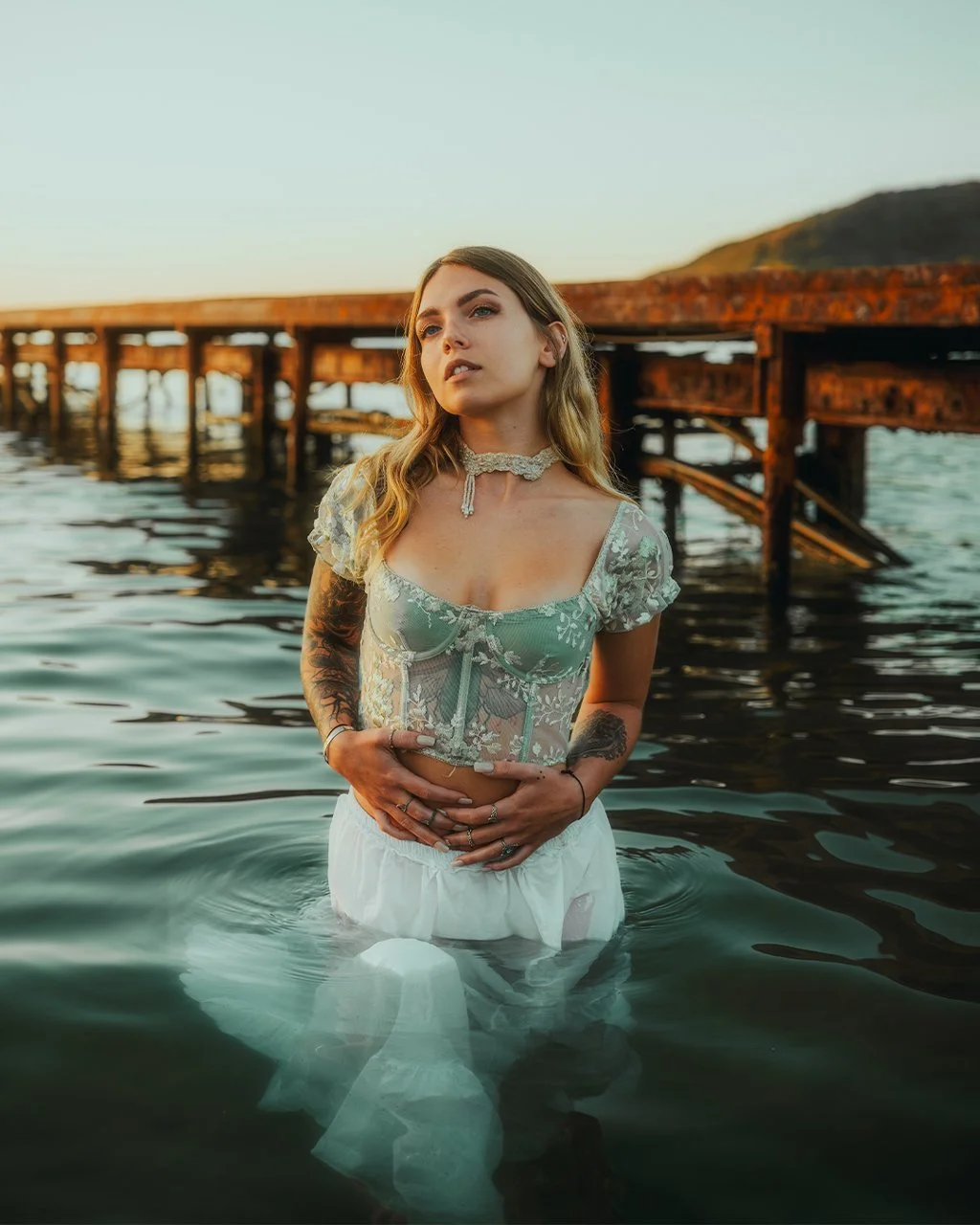 Jeune femme aux tatouages debout dans l'eau près d'une jetée en bois, portant une robe en dentelle verte et une choker, au coucher du soleil.