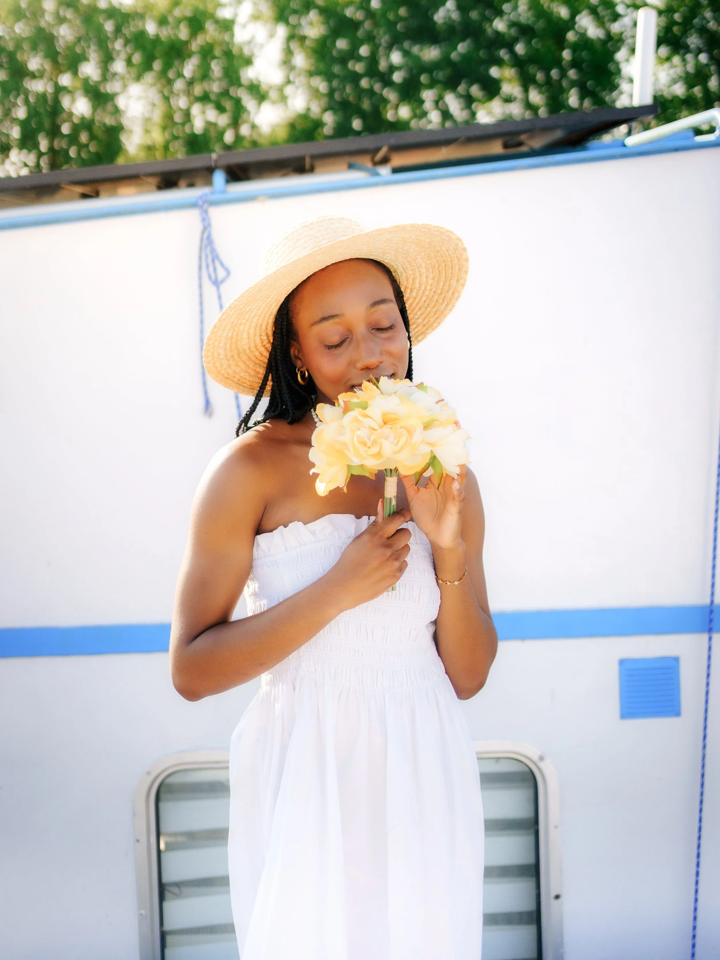 Jeune femme portant un chapeau en paille blanc, vêtue d'une robe blanche, tenant un bouquet de fleurs crème, en extérieur avec un mur blanc et des arbres en arrière-plan.