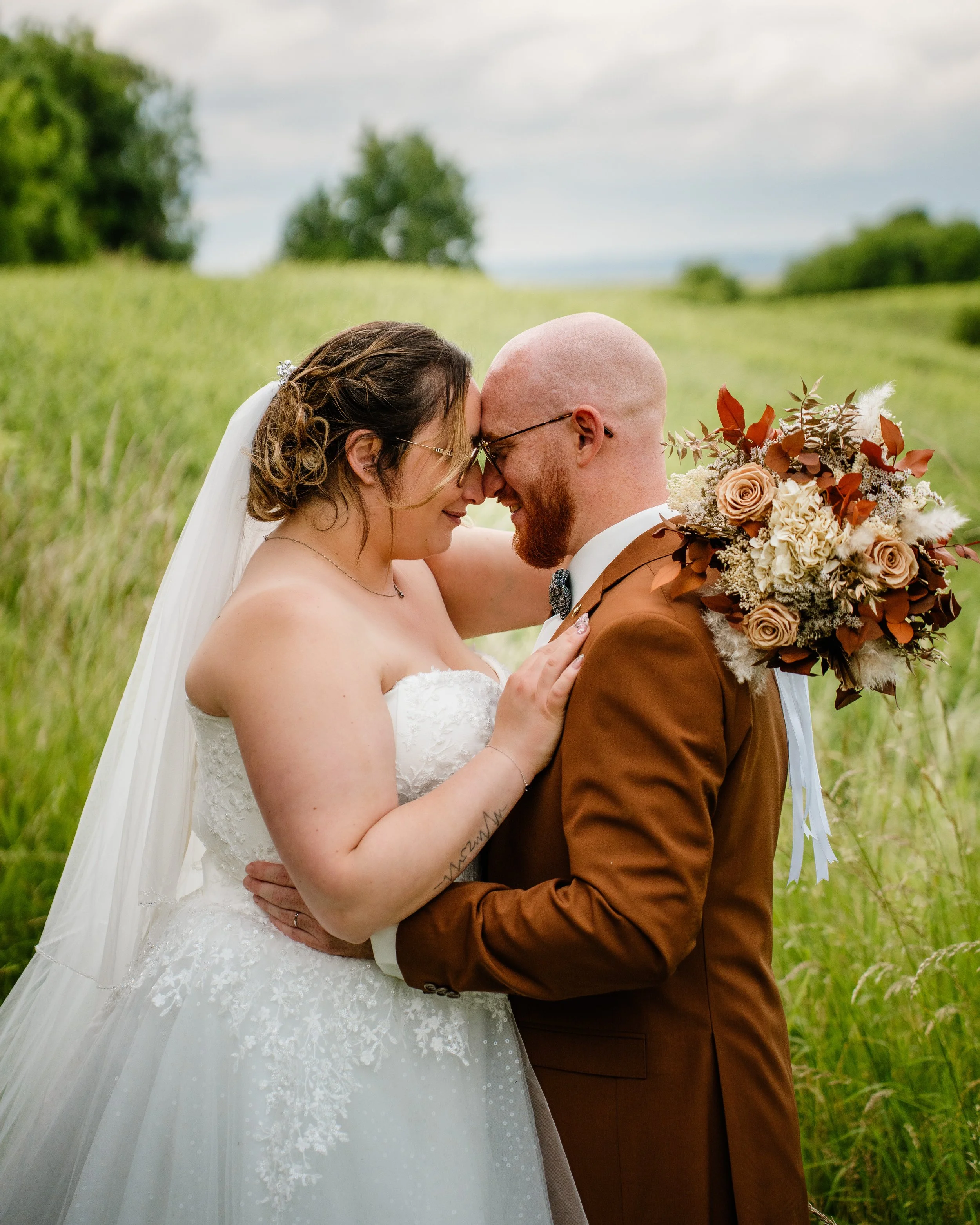 Un couple de mariés s'embrassant dans un champ, lors d'une séance photo de mariage, la femme porte une robe blanche et le homme un costume marron, avec un bouquet de fleurs.