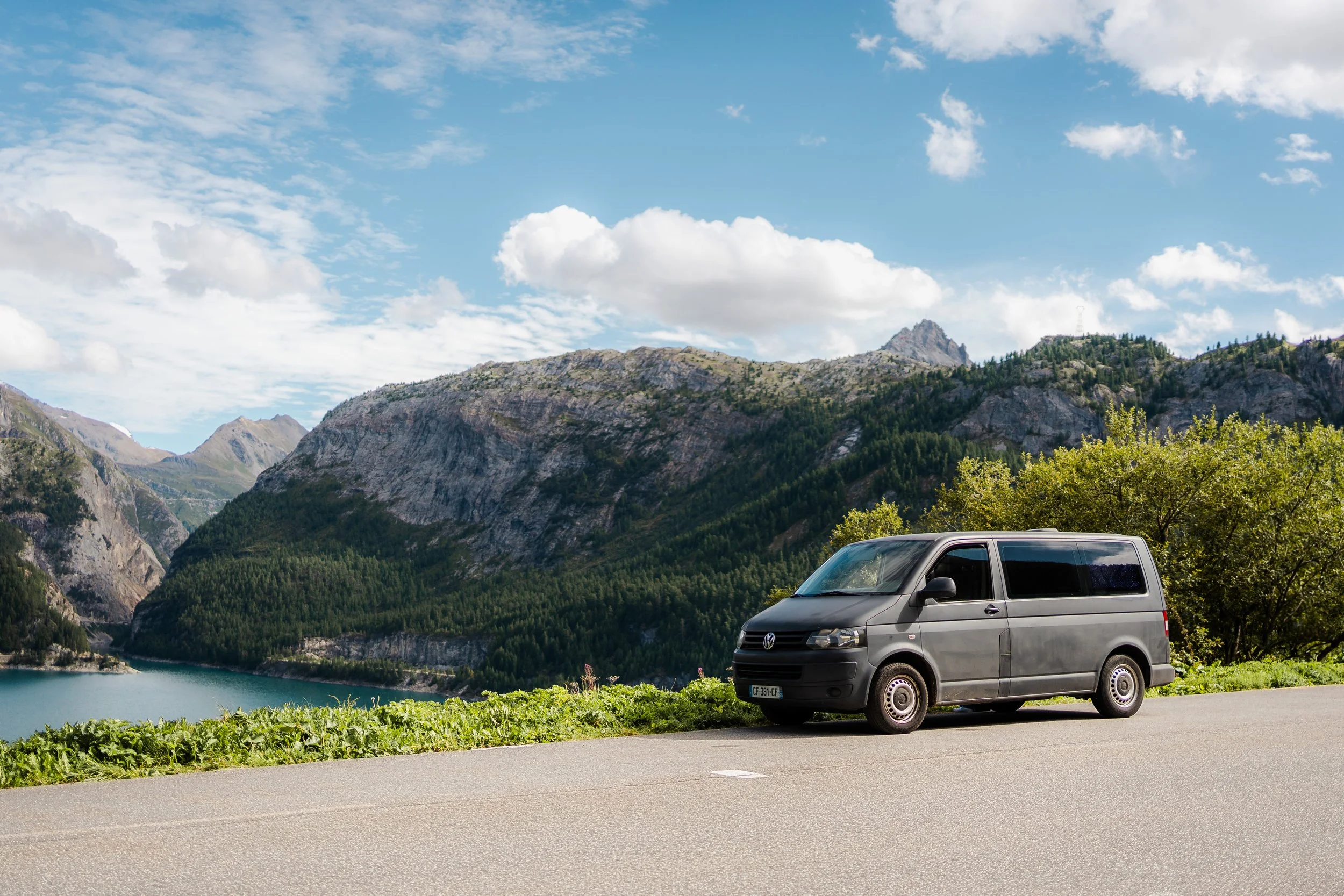 Une voiture noire garée sur le côté d'une route au bord d'un lac entouré de montagnes, avec un ciel bleu et quelques nuages.