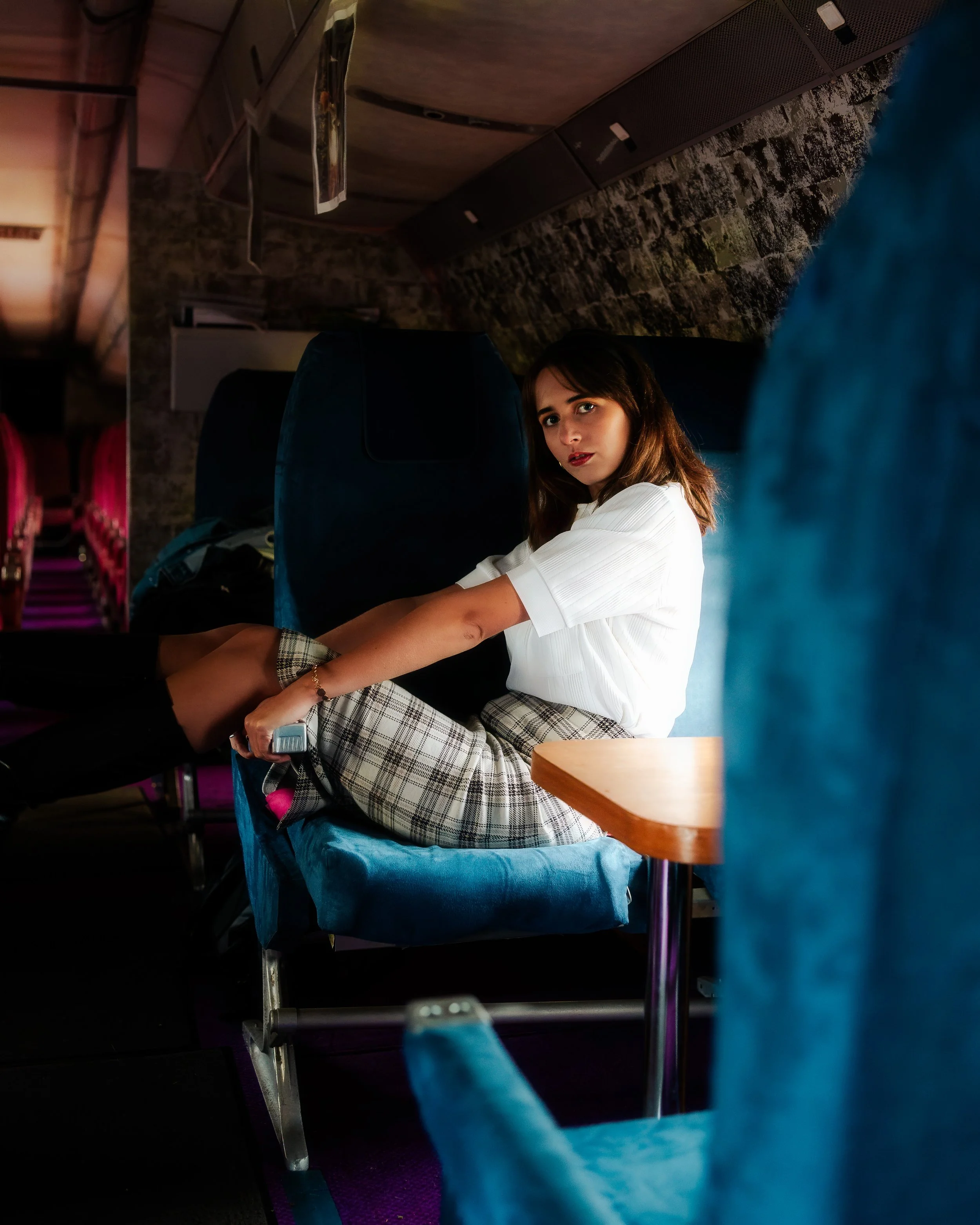 Une jeune femme assise sur une banquette dans un bus ou un train, portant un chemisier blanc et une jupe à carreaux, regardant vers la caméra.