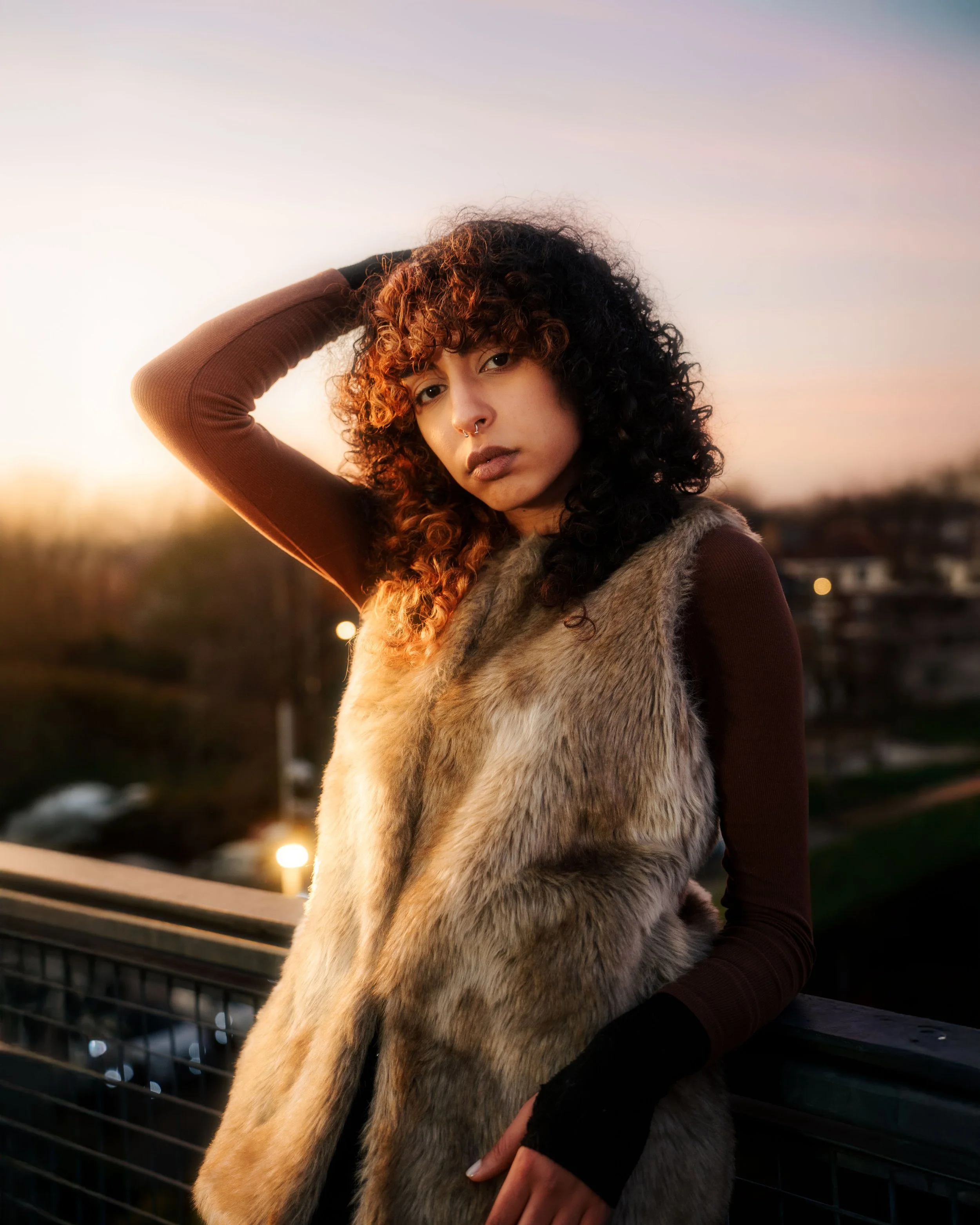 Une jeune femme aux cheveux bouclés avec des mèches colorées, portant un gilet en fourrure et un haut à manches longues, se tient sur un pont au coucher du soleil.
