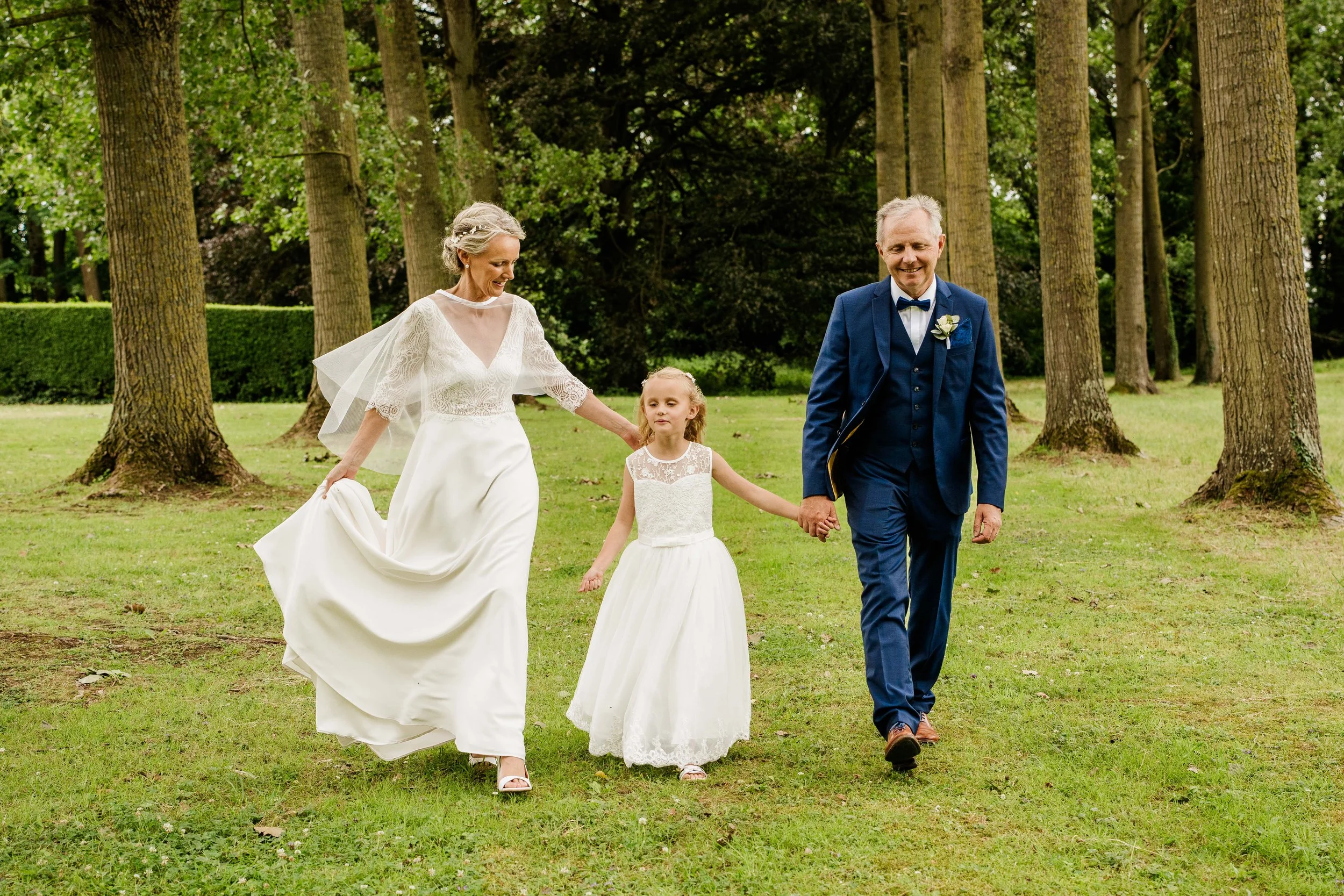 Une famille en costume de mariage marchant dans un parc avec des arbres en arrière-plan, la mariée en robe blanche, l'homme en costume bleu, une jeune fille en robe blanche.