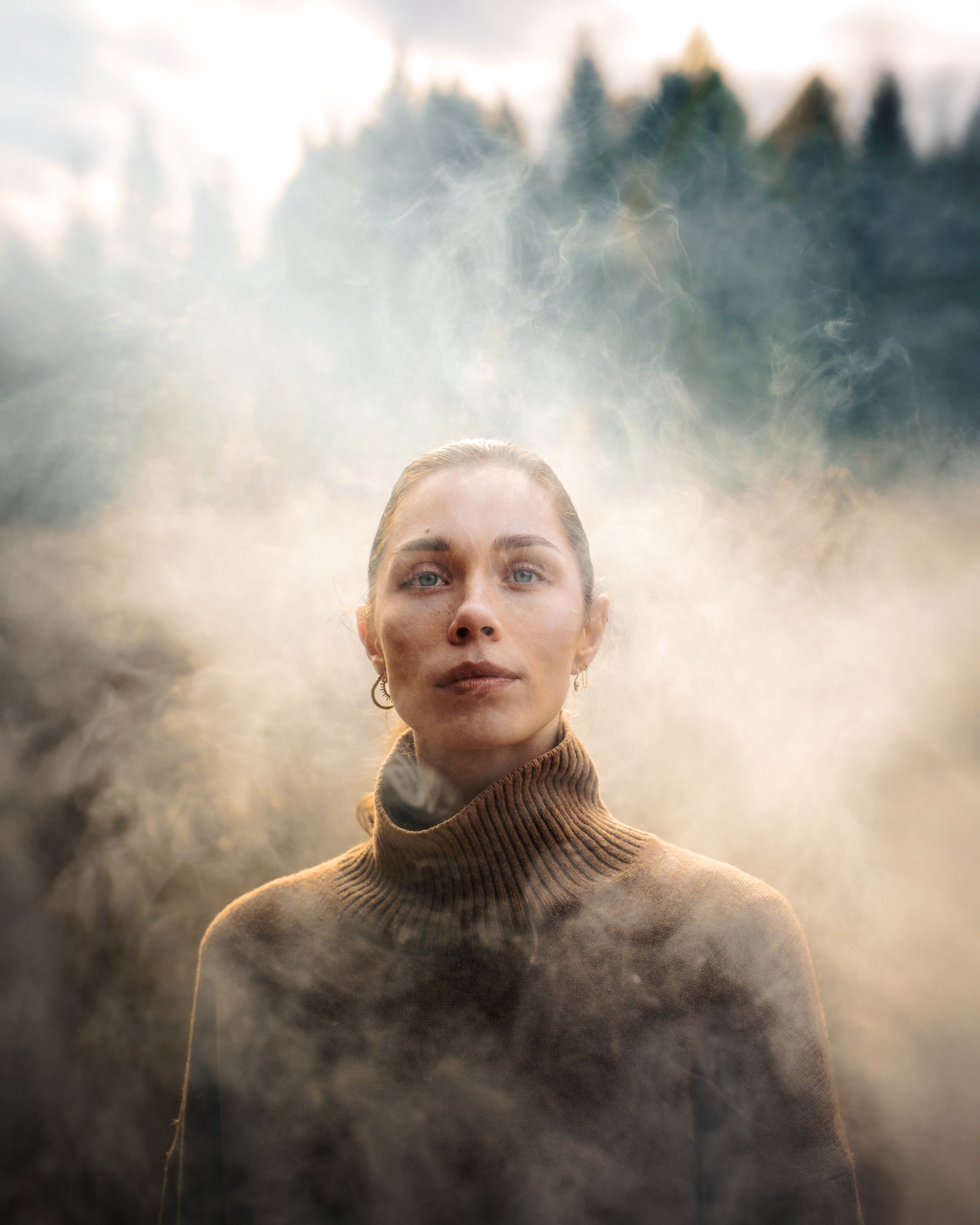 Femme en pull marron clair, entourée de vapeur ou de brouillard, avec un paysage forestier en arrière-plan.