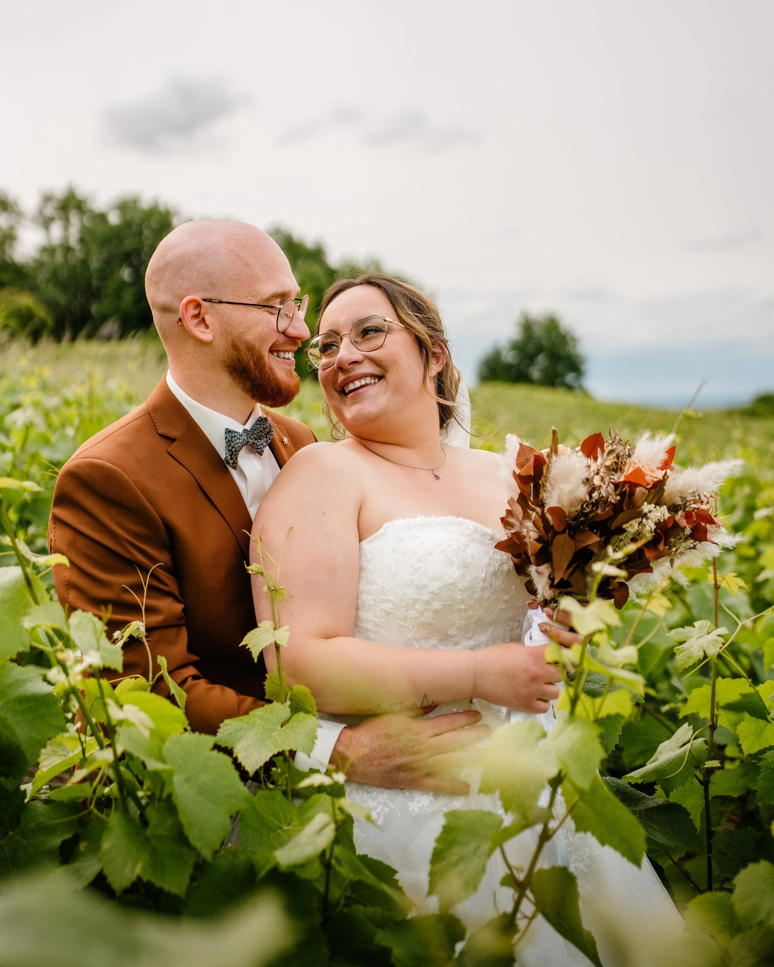 Un couple de mariés souriants dans un champ de vignes, la femme tenant un bouquet de fleurs, le marié en costume marron. Ciel nuageux en arrière-plan.