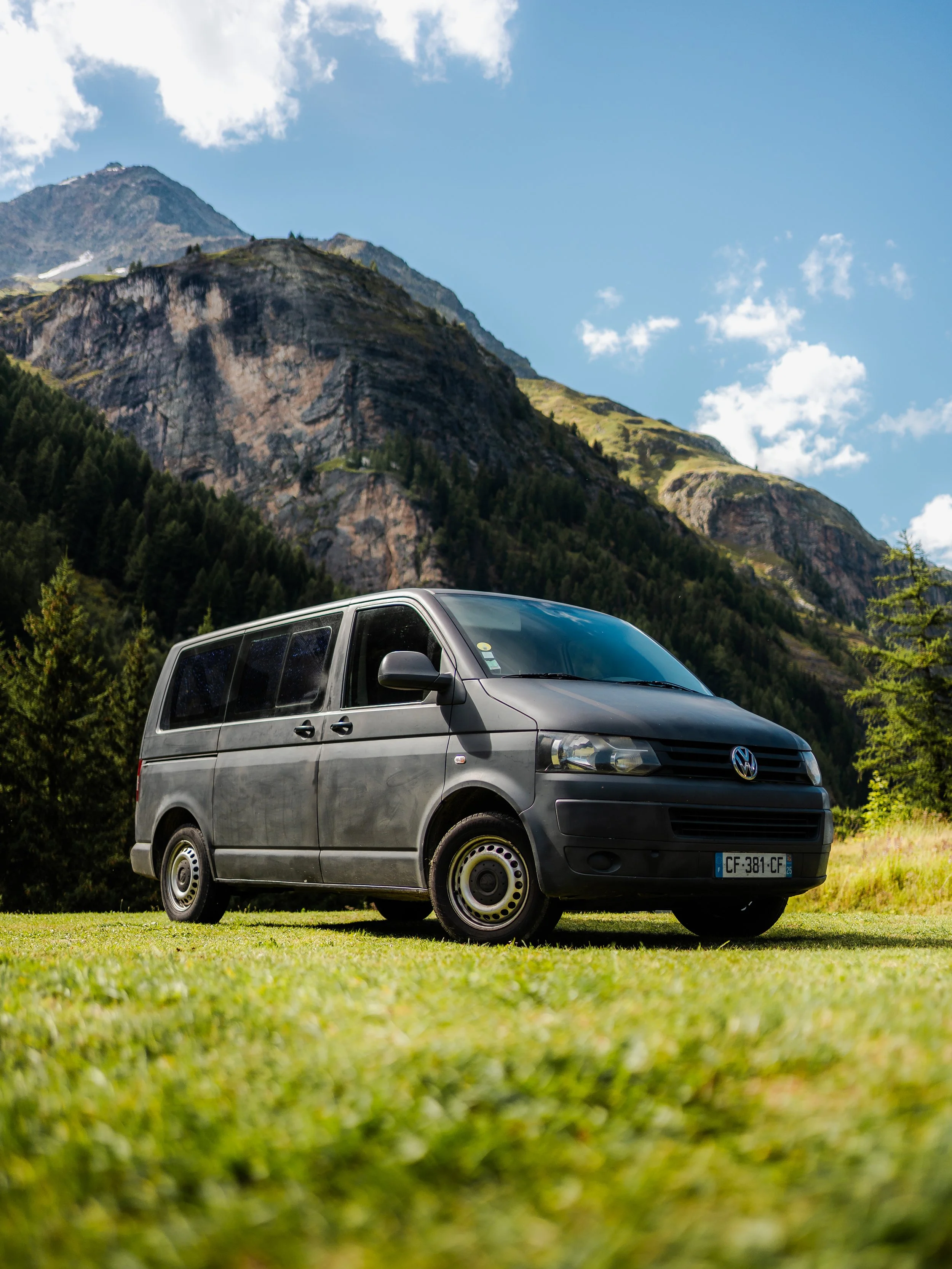 Une voiture noire Volkswagen garée sur une prairie verte avec des montagnes en arrière-plan et un ciel bleu avec quelques nuages.