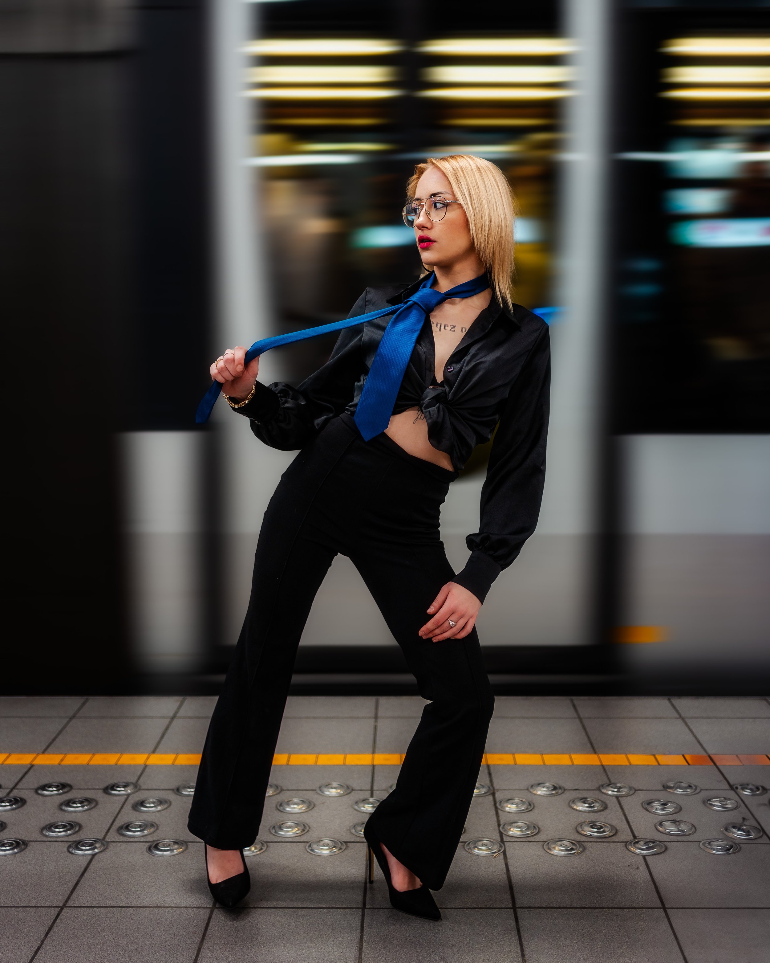 Une jeune femme élégante porte une robe noire satinée, un nœud en cravate bleu autour du cou, et des talons noirs, posant sur un quai de métro ou de train avec un train en mouvement en arrière-plan.