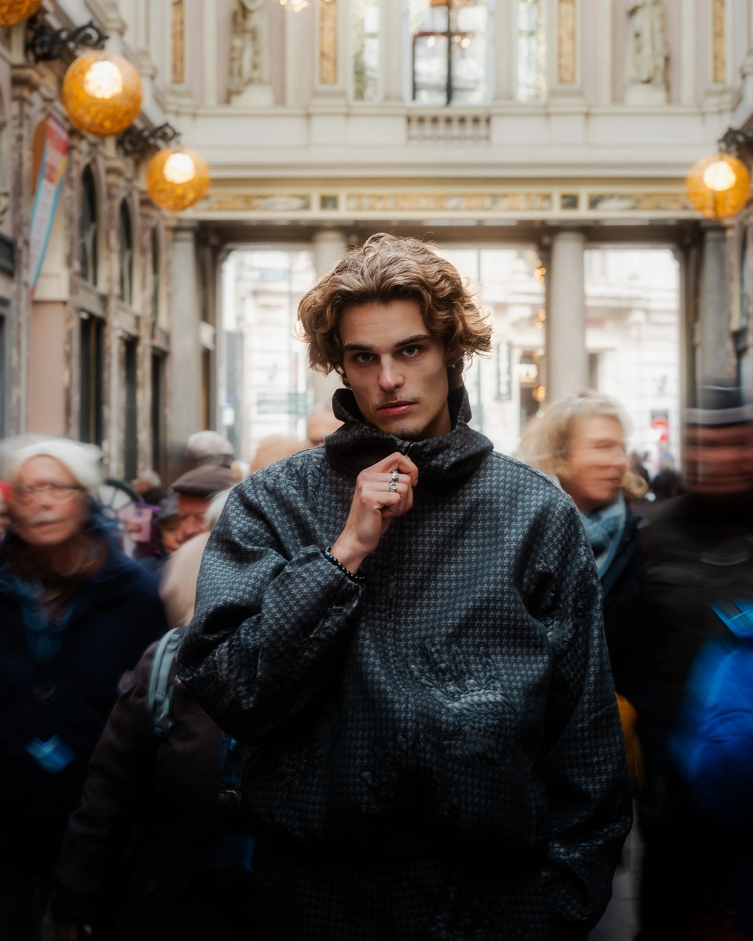 Un jeune homme avec des cheveux bouclés portant une veste sombre dans une galerie commerçante animée, entouré de personnes floues.