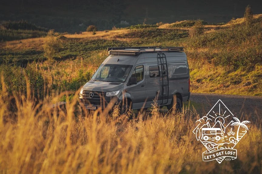 Ein grauer Campervan steht auf einer Landstraße in einer ländlichen Umgebung bei Sonnenuntergang, umgeben von Gras und Bäumen.