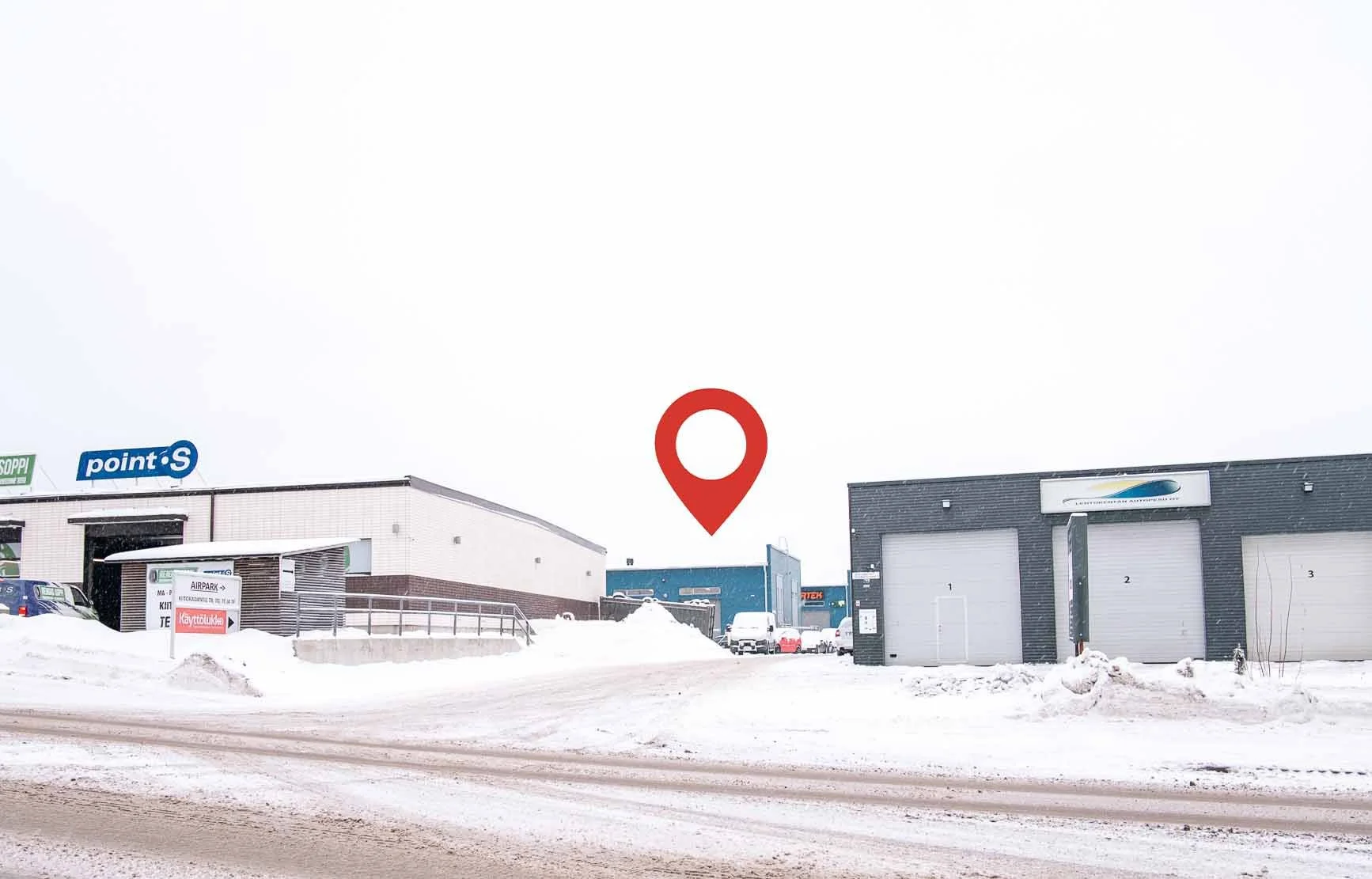 Luminen ja tatoilla päällystetty tie johtaa teollisuusalueelle, jossa on varastoja ja pysäköintialueita, ympärillä lumimonttuja ja rakennuksia, taivaalla harmaa pilviverho.