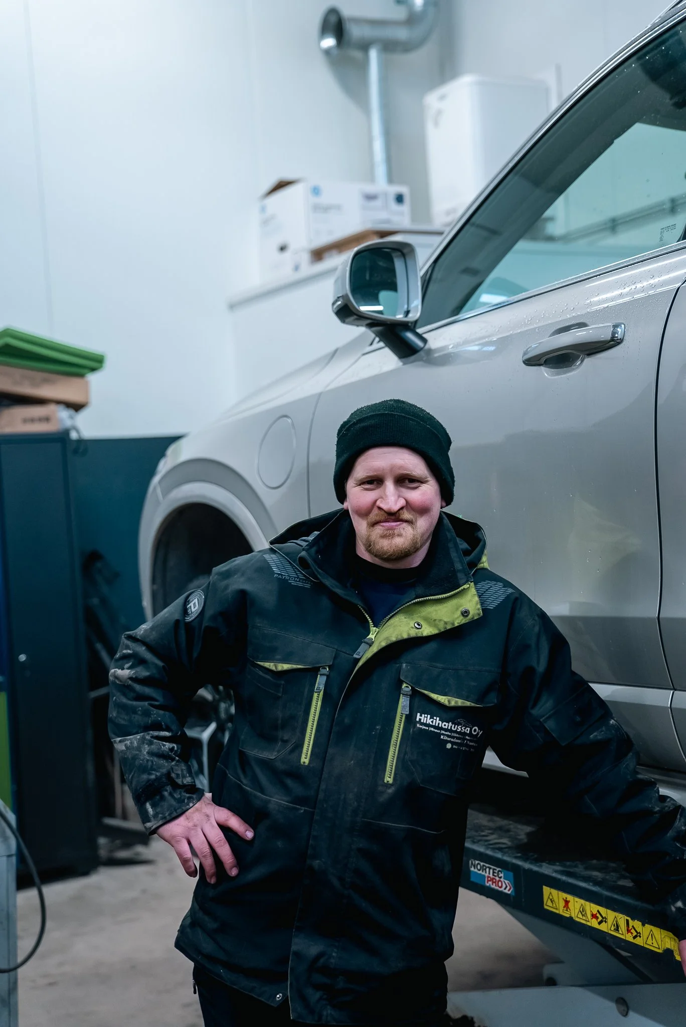 Mies poseeraa autotallissa talvitakin ja pipo päässään, vieressään autotalliostinlaitteisto ja auto.