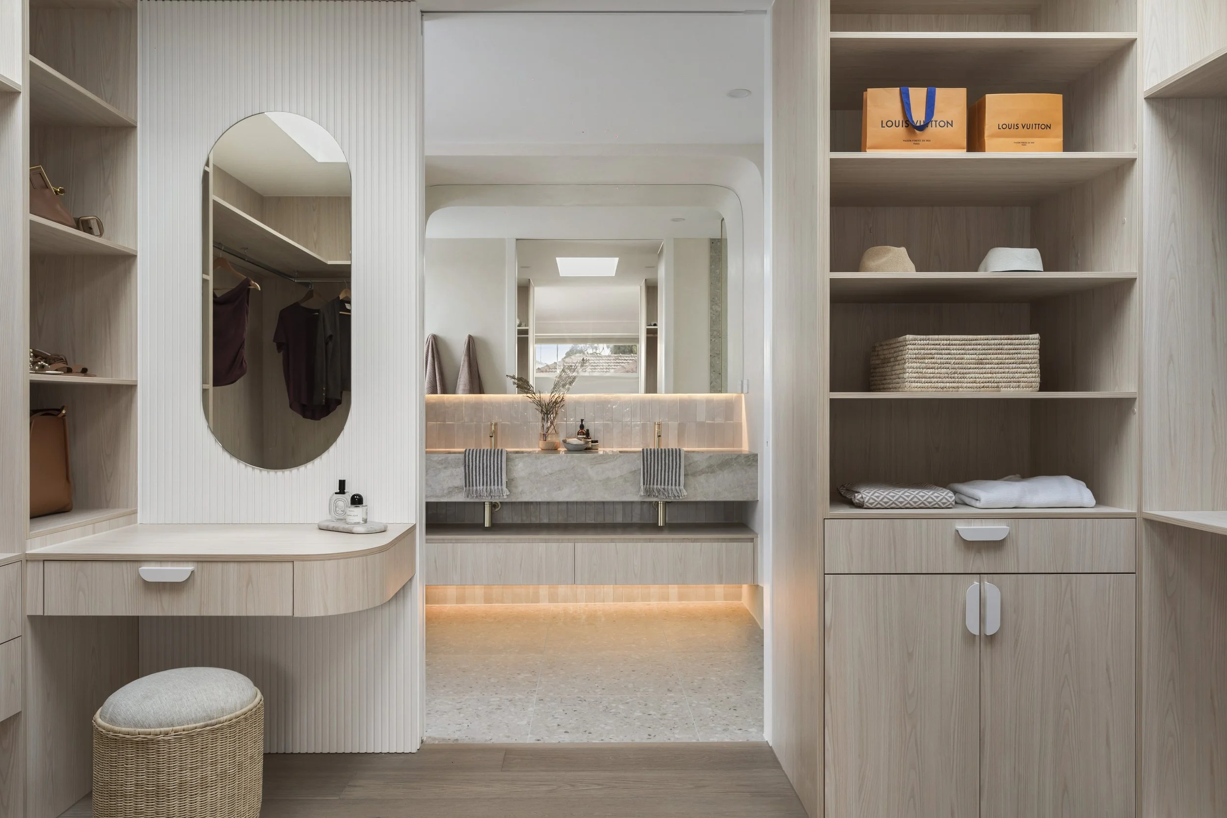 Modern bathroom with a vanity mirror, shelves with handbags, hats, towels, and drawers, soft lighting, and a window view in the background.