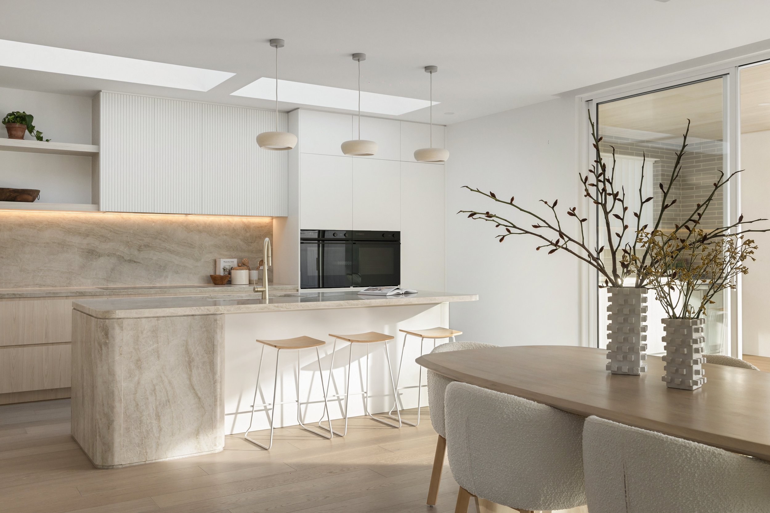 Modern, minimalist kitchen with white cabinetry, marble countertops, and a breakfast bar with three bar stools. There are three pendant lights hanging from the ceiling and a large sliding glass door. On the dining table, there are two vases with bran