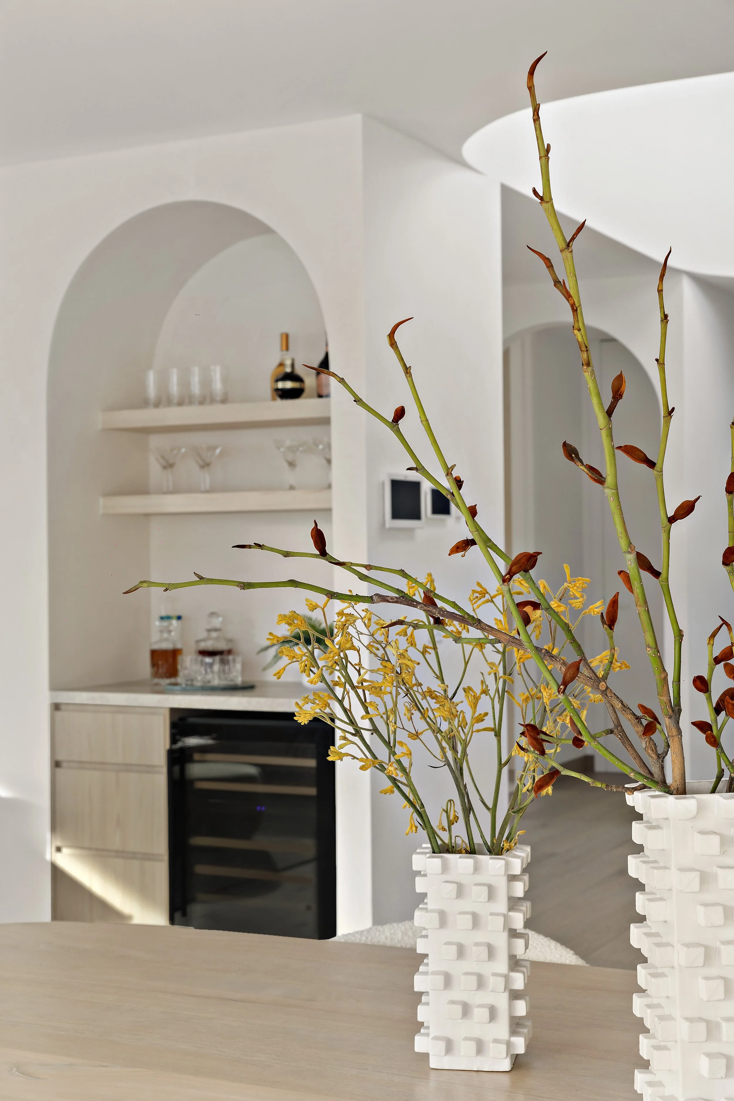 Two white textured vases with dried branches and yellow flowers on a wooden table in a modern, minimalist kitchen.