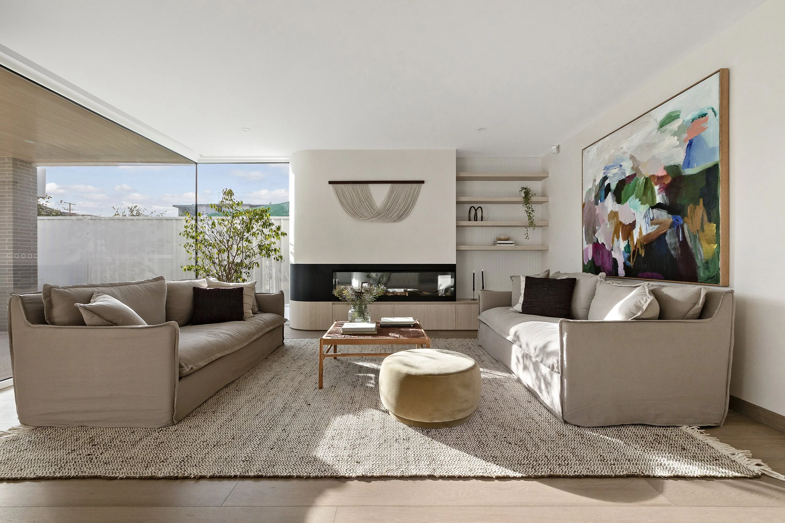 Modern living room with two beige sofas, a wooden coffee table, a plush ottoman, a large abstract painting, and built-in shelves with decorative items. Natural light coming through large windows with a view of the outdoors.