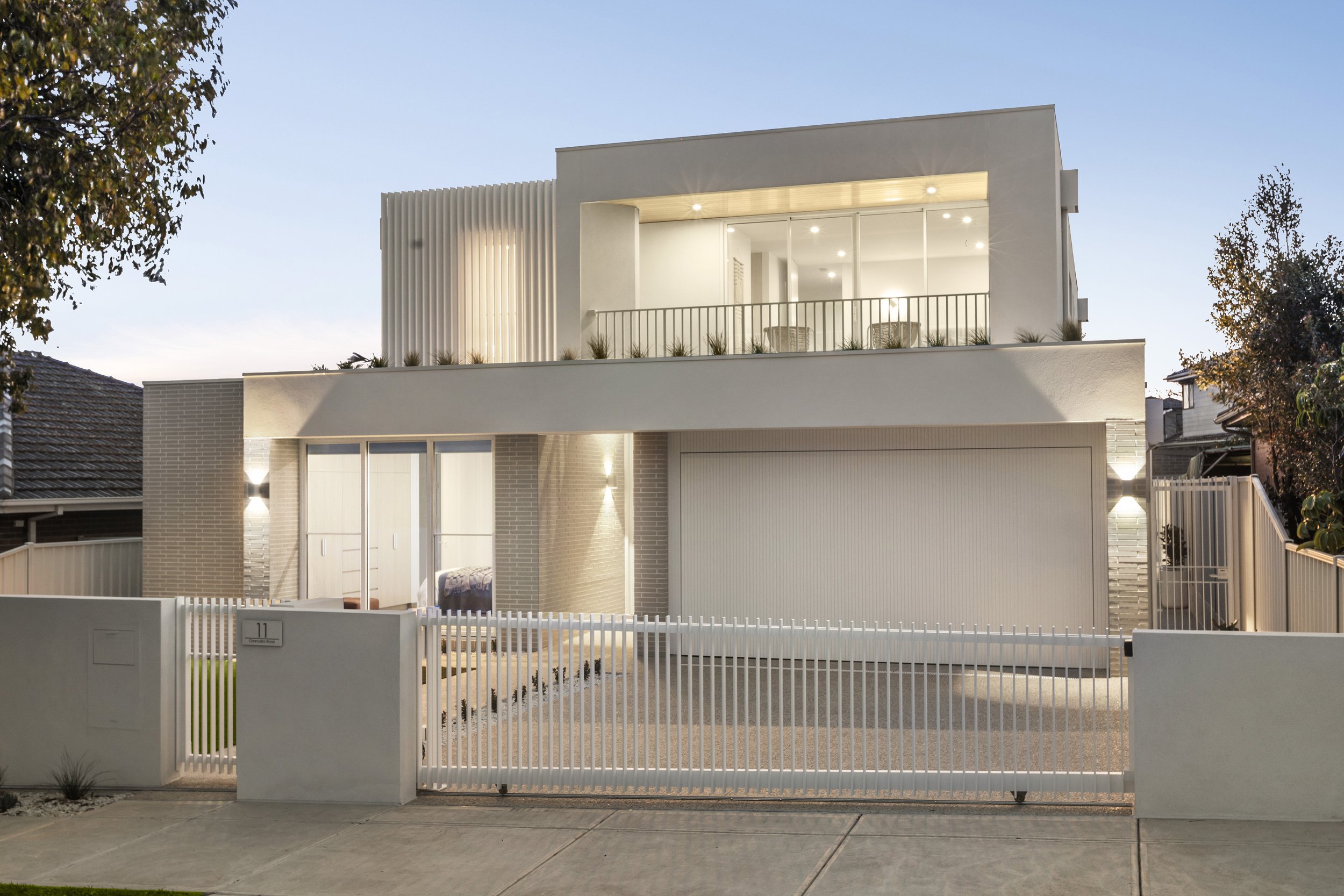 Modern two-story house with white exterior, large glass doors, and a white fence, illuminated at dusk with exterior lights.