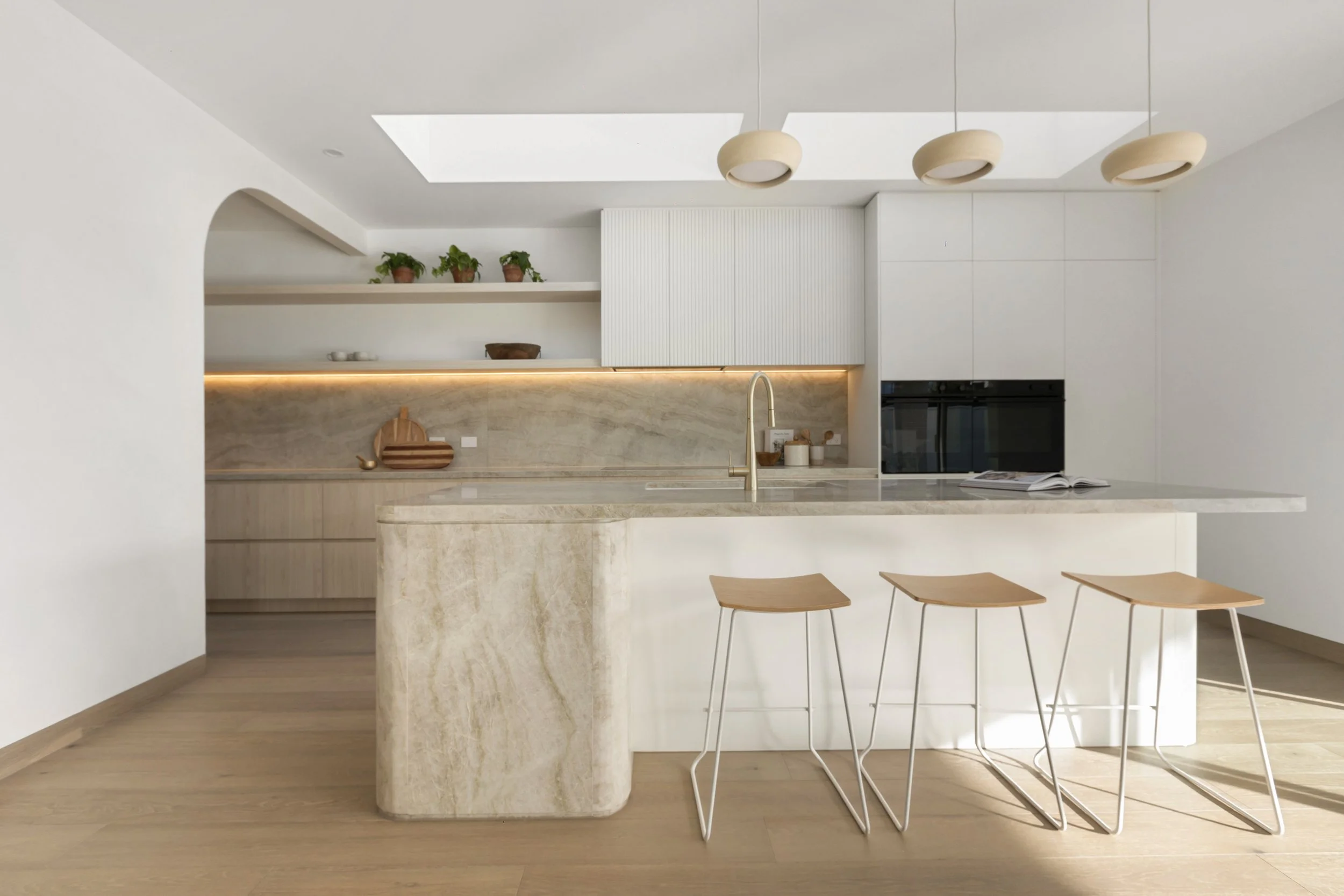 Modern kitchen with white cabinets, light wood bar stools, marble island, open shelving with plants, illuminated backsplash, built-in oven, and minimalist design.