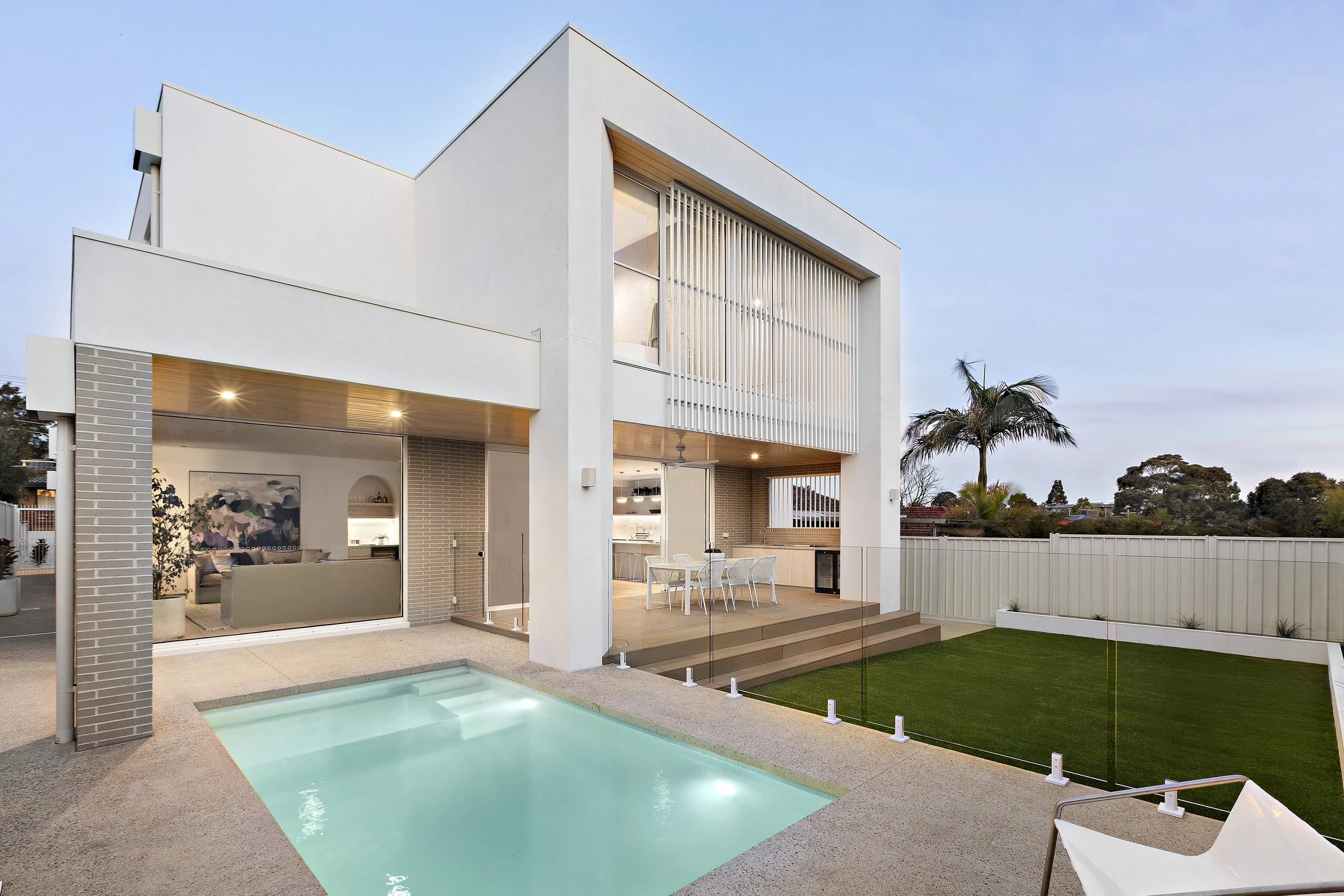 Modern white house with a backyard featuring a swimming pool, outdoor dining area, and a green lawn, with a tall palm tree and a white fence.