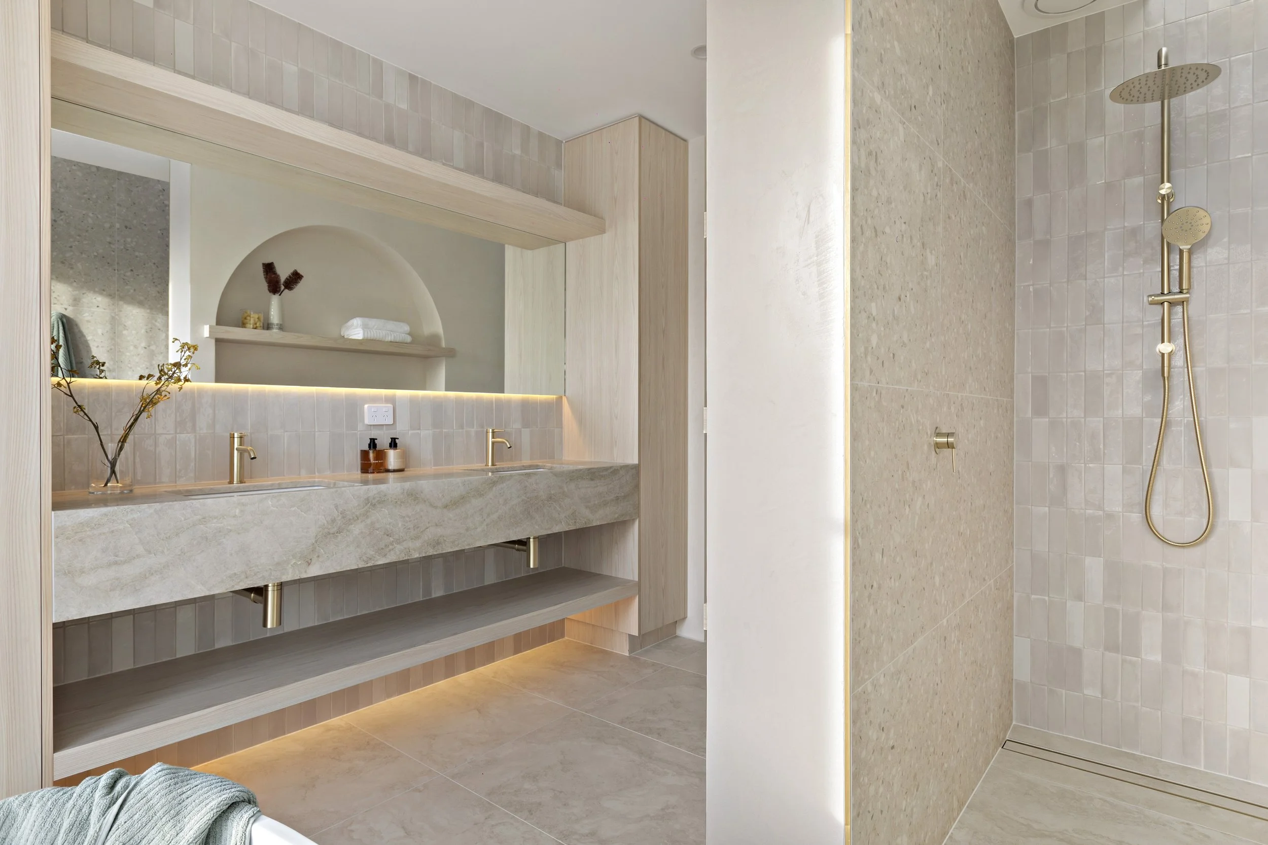Modern bathroom with double vanity, beige marble countertop, gold fixtures, and walk-in shower with beige tiles and wall-mounted showerhead