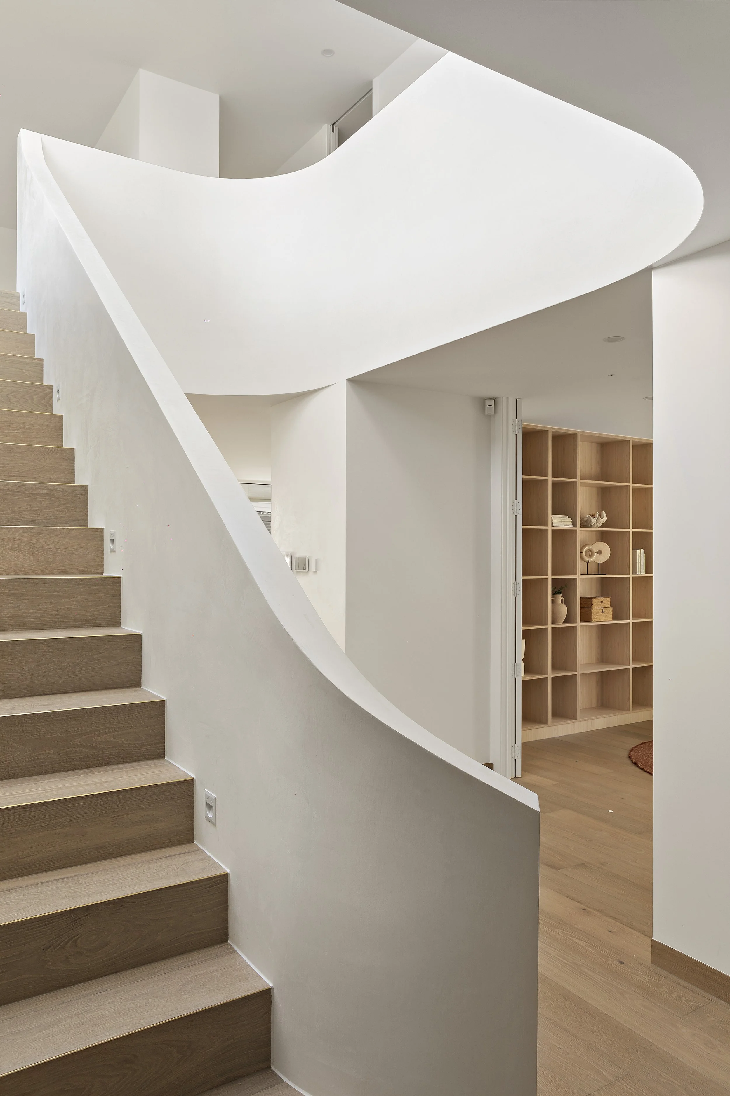 Interior of a modern home featuring a wooden staircase, a curved white wall, and a wooden shelving unit with decorative items.