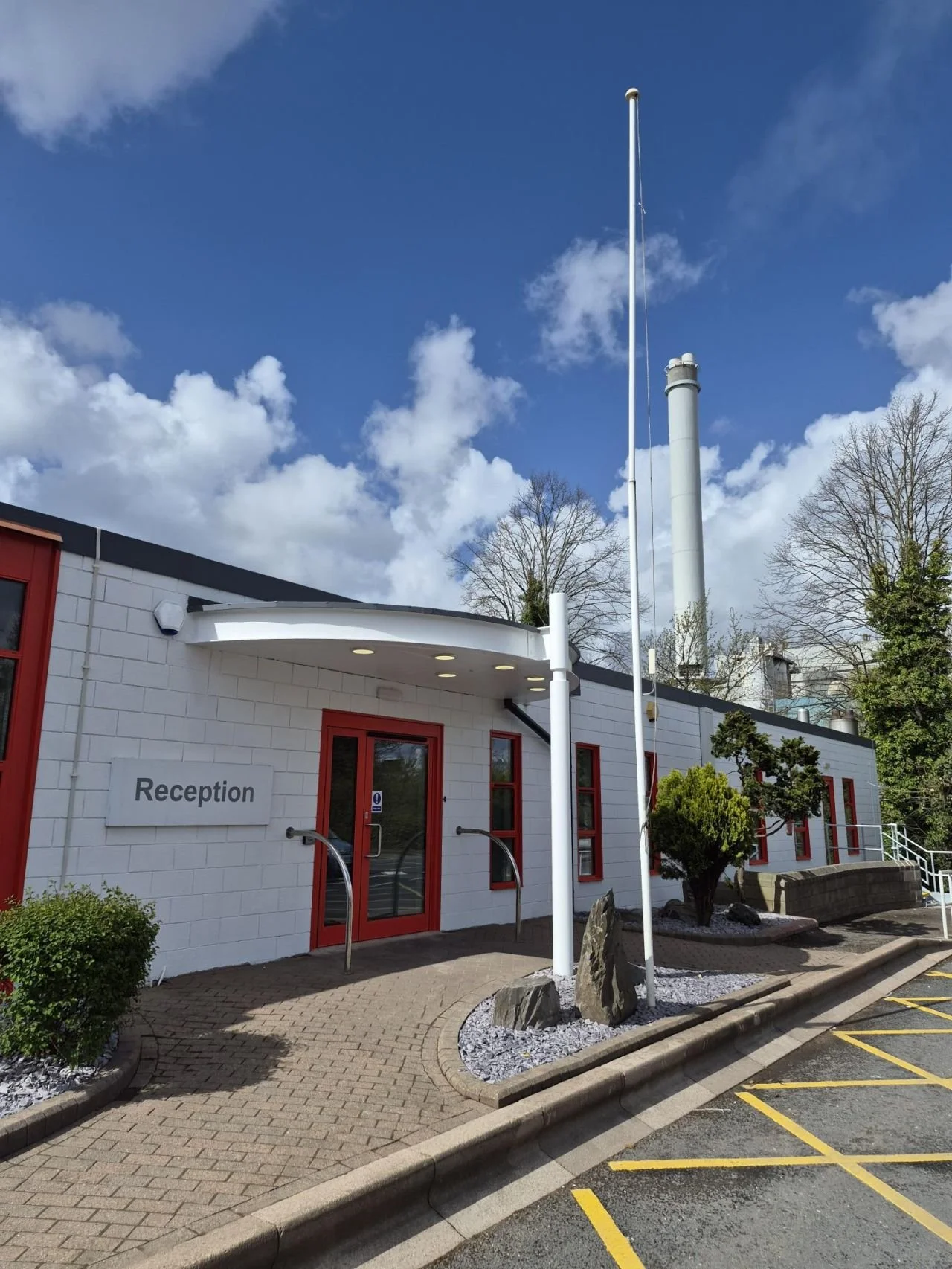 Hospital or clinic reception building with white brick walls, red trim, and a sign that says 'Reception'. There are plants, rocks, and a paved walkway in front, with a flagpole and smoke stacks in the background under a partly cloudy blue sky.