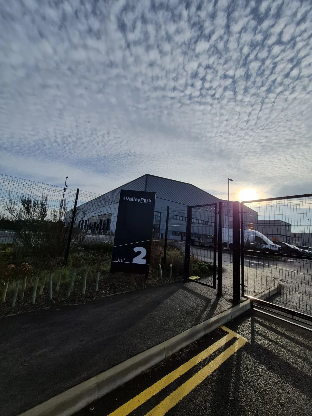 The image shows a modern commercial or industrial building with a sign that reads 'ValleyPark, Unit 2'. The building is behind a black metal fence, and the sun is setting or rising in the background, casting a bright light and creating shadows. The sky is partly cloudy with a textured pattern of clouds.