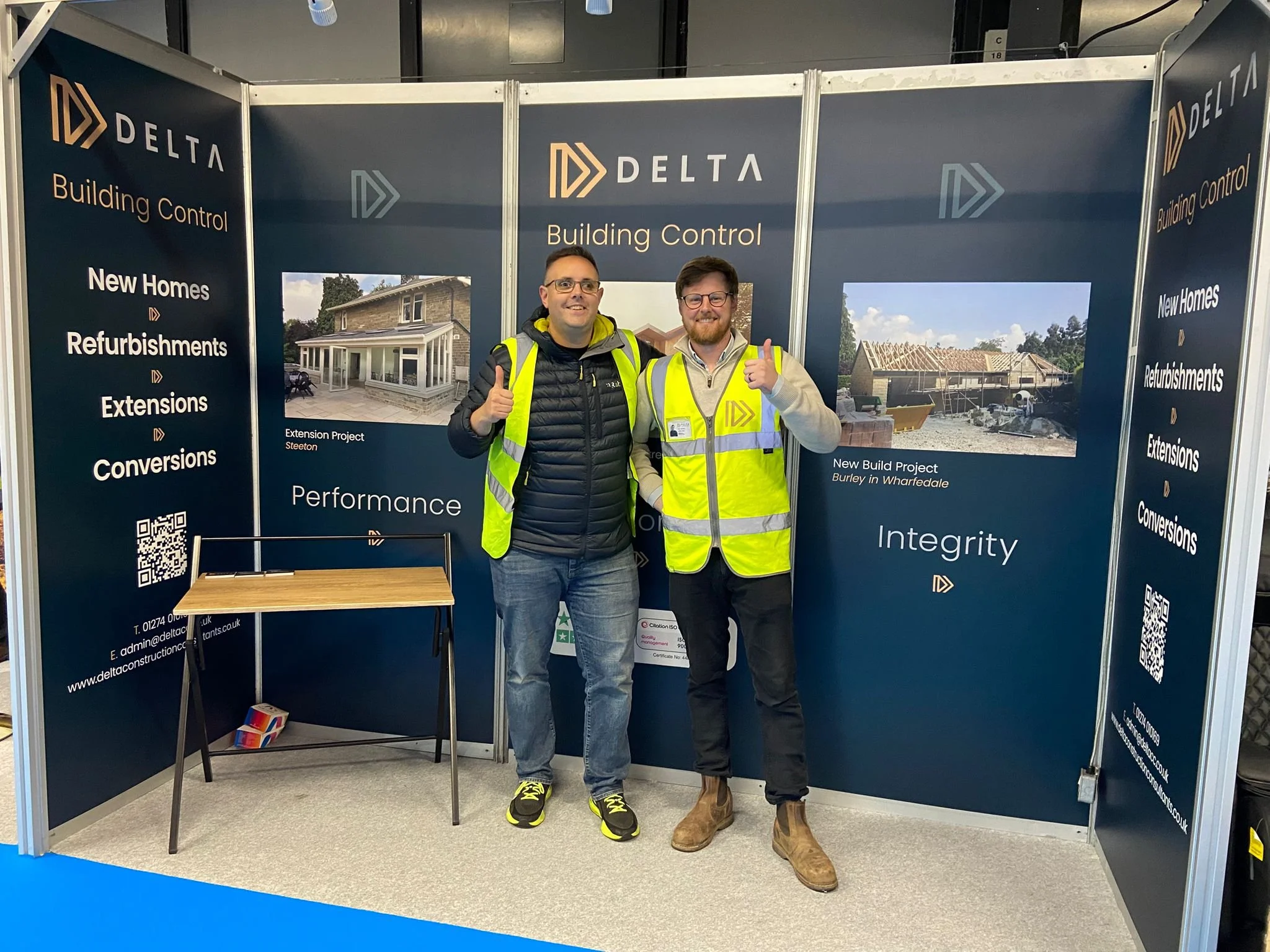The company directors standing inside a Delta Building Control booth at an exhibition, smiling and giving thumbs-up gestures. They are dressed in casual and safety gear, with the booth displaying information about building projects and services.