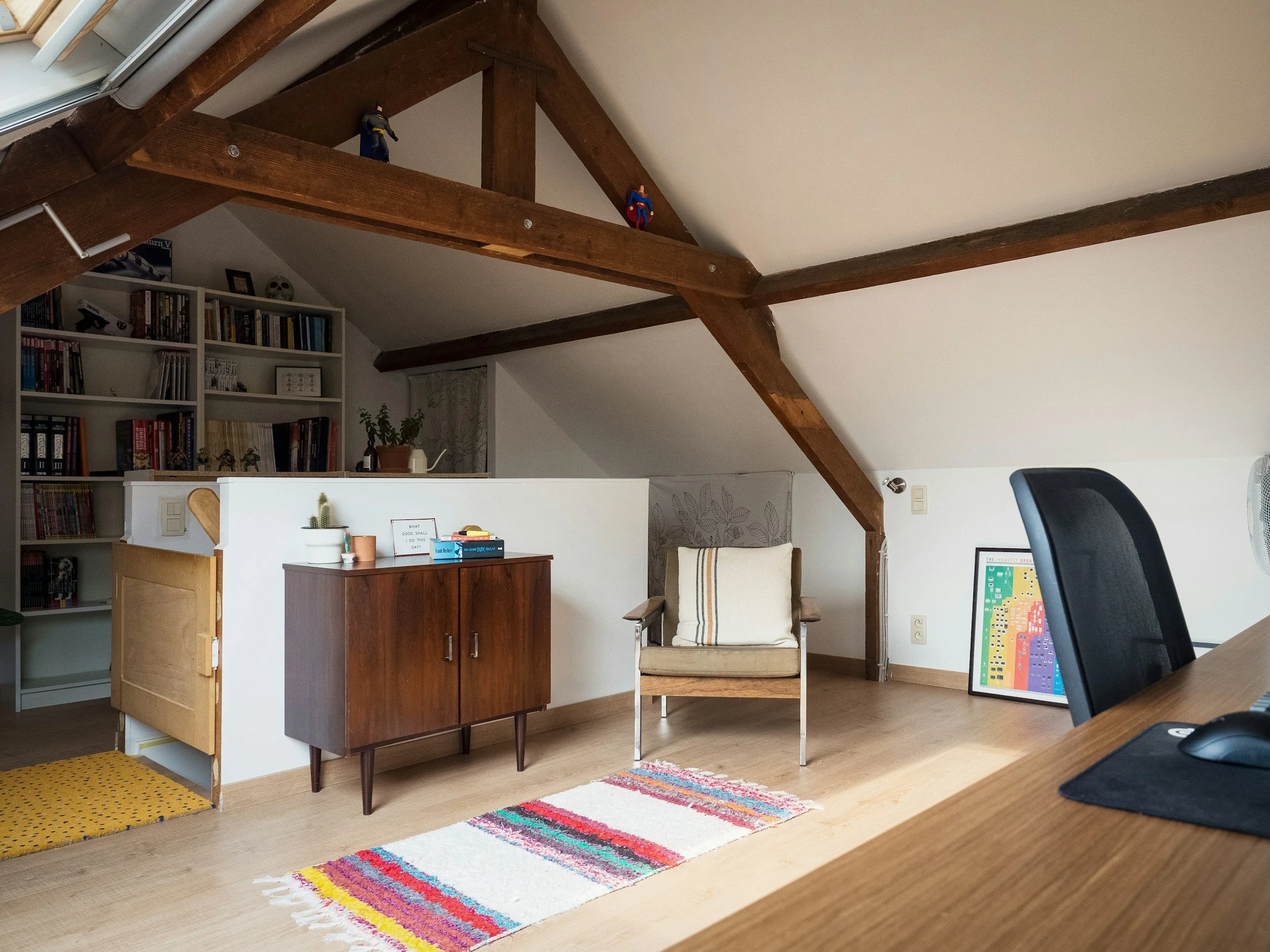 Living room with exposed wooden beams, bookshelf, armchair, colorful striped rug, and a framed colorful artwork on the floor