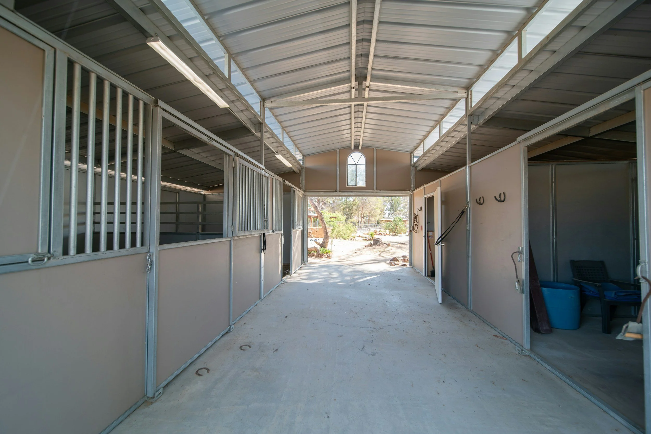 Interior of a metal-framed barn with stalls, high arched ceiling, and natural light through windows and open doors, overlooking an outdoor area.