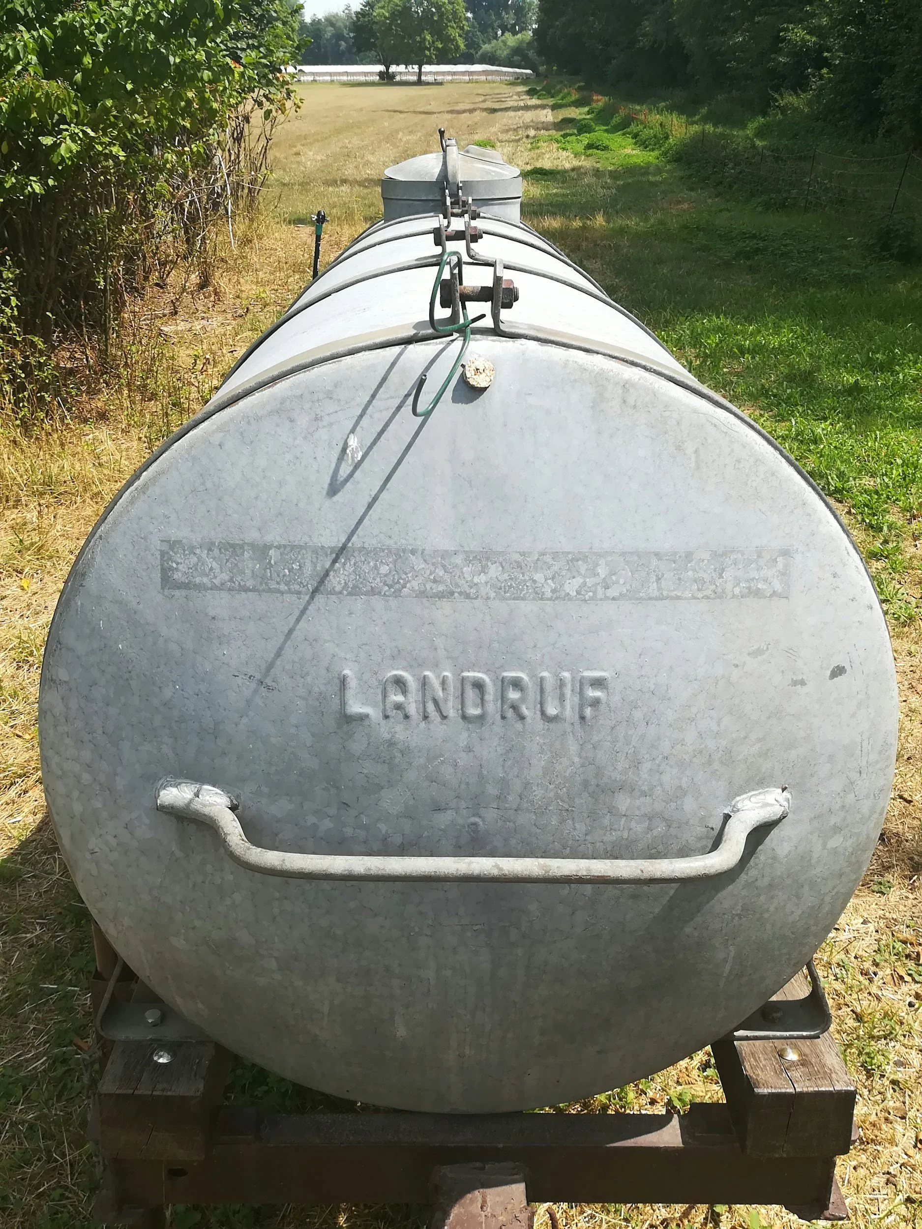 A metal water tank with the word 'LANDRUFF' embossed on it, placed on a wooden pallet outdoors, with trees and green grass in the background.