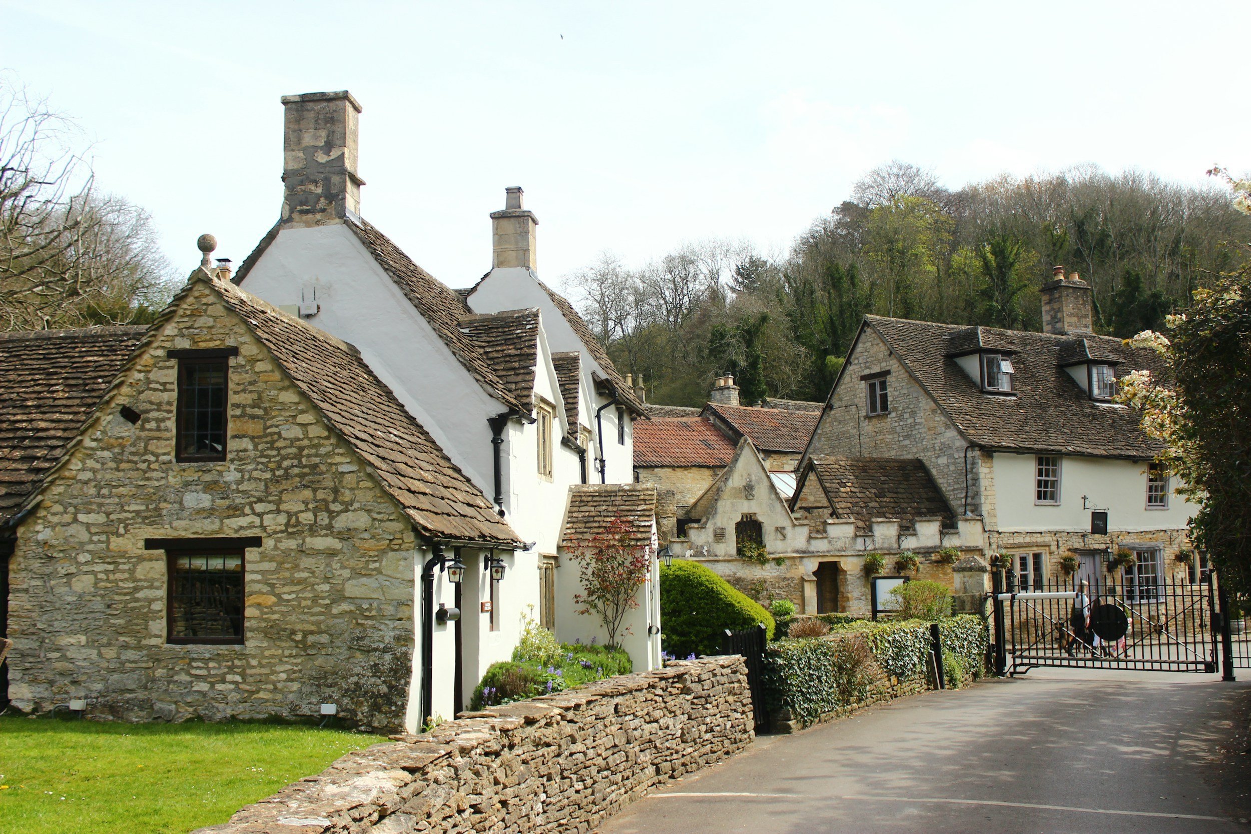 A row of stone and white cottages with pitched roofs and chimneys in a scenic village setting, with a cobblestone wall and a gate in the foreground and trees on a hill in the background.