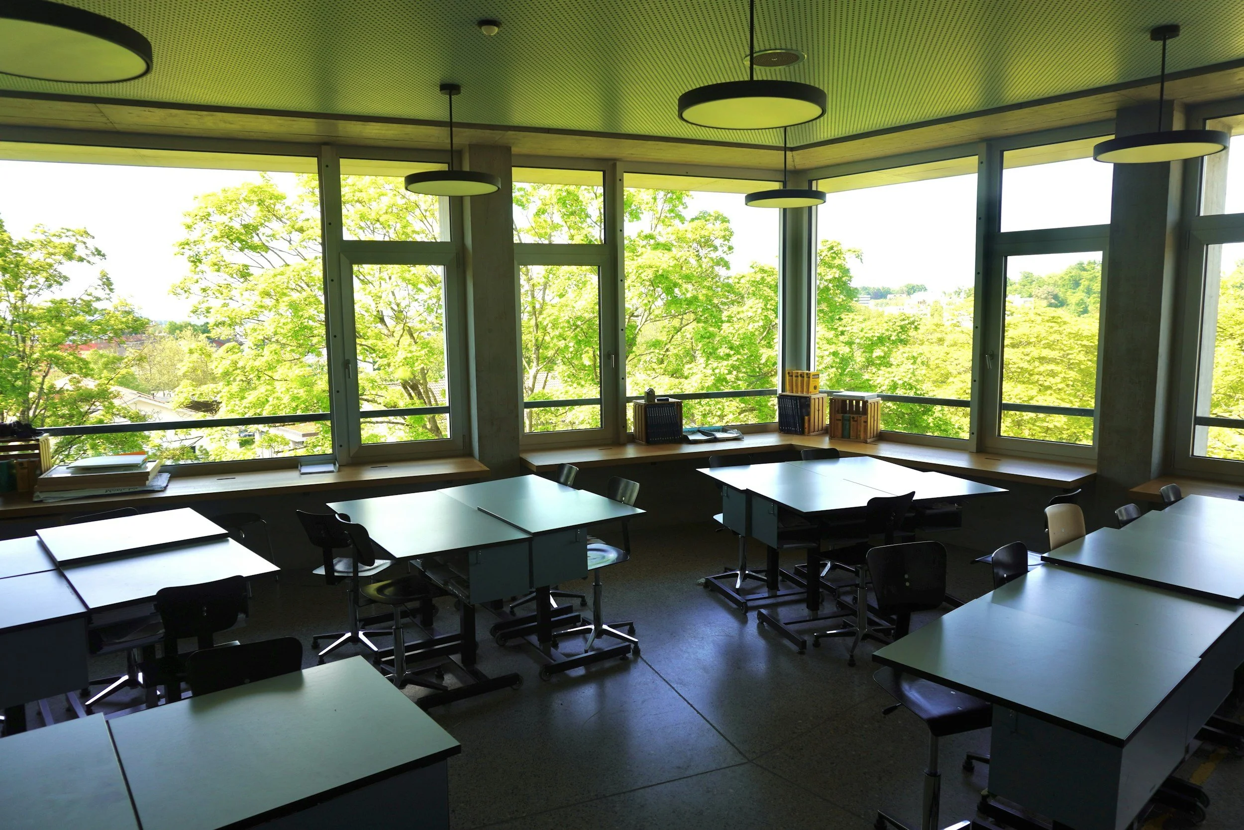 A modern conference room with large floor-to-ceiling windows showing green trees outside. There are tables and chairs arranged for a meeting, with books and documents on the tables and window sill. The ceiling has circular pendant lights.