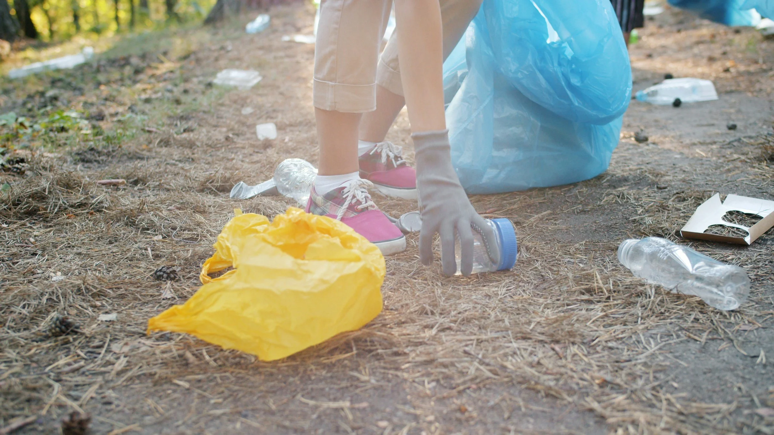 Person wearing beige shorts, plaid shoes, and gloves picking up trash, including plastic bottles, in a wooded area during the day, with a large blue trash bag nearby and litter scattered on the ground.