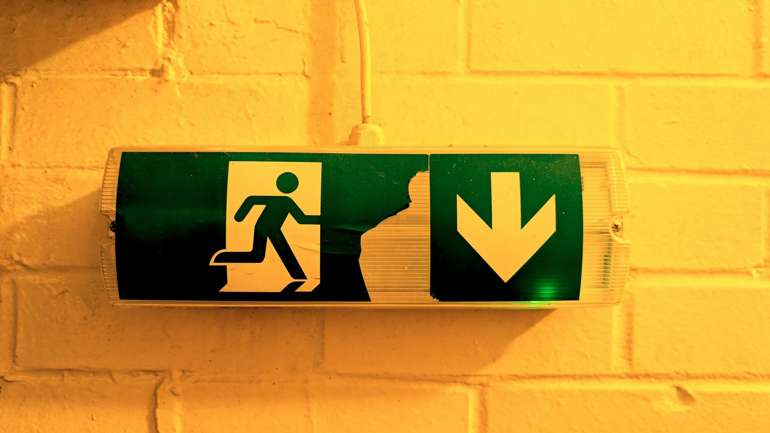 Emergency exit sign with a running person figure and an arrow pointing downward, mounted on a yellow brick wall.